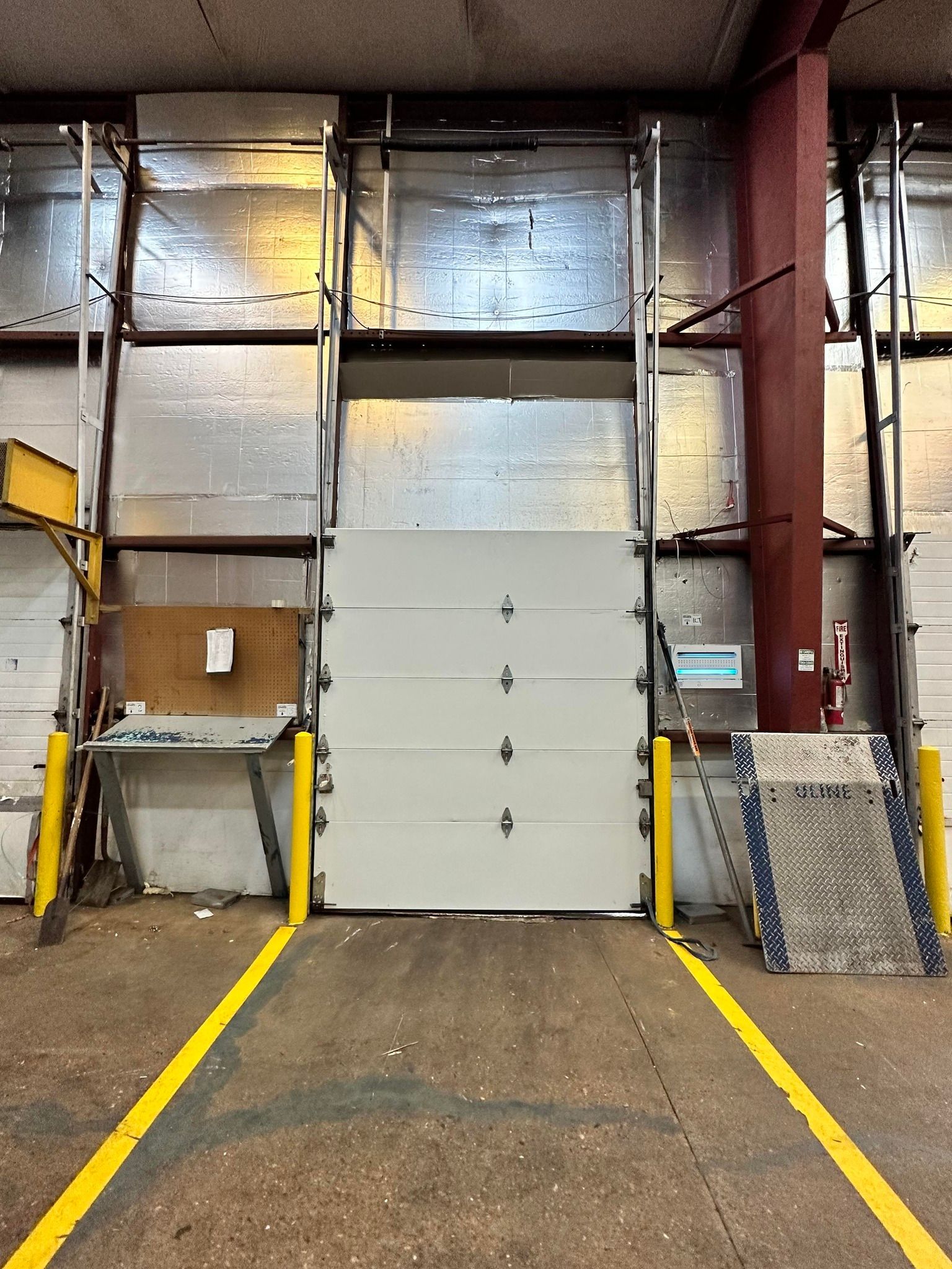 A loading dock with a closed white sectional door, framed by yellow safety bollards and floor markings inside a warehouse.