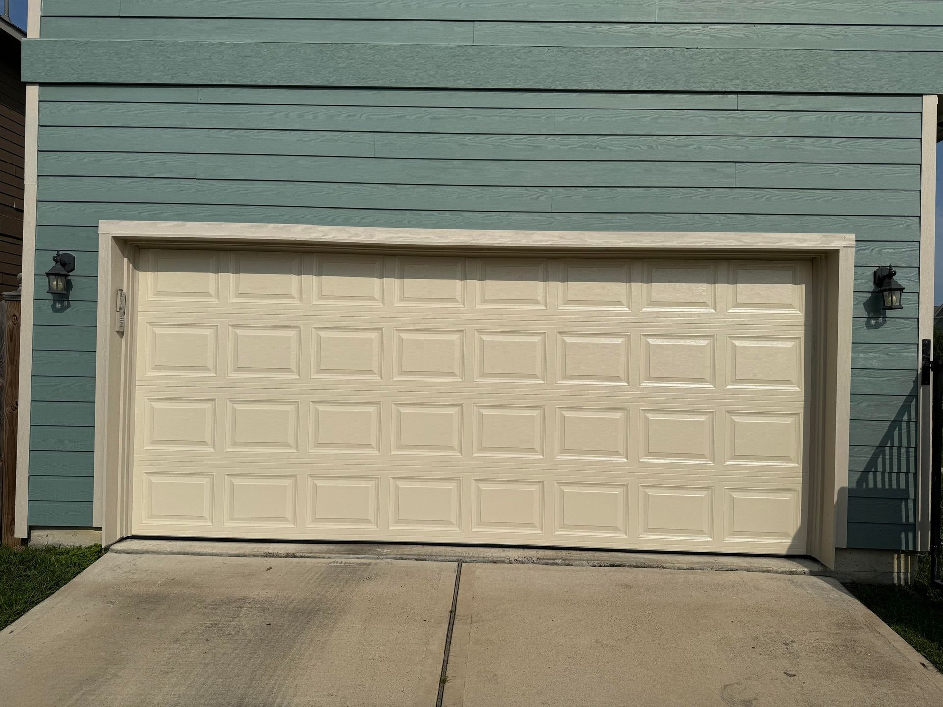 A cream-colored, multi-panel garage door installed on a house with blue horizontal siding and a concrete driveway.