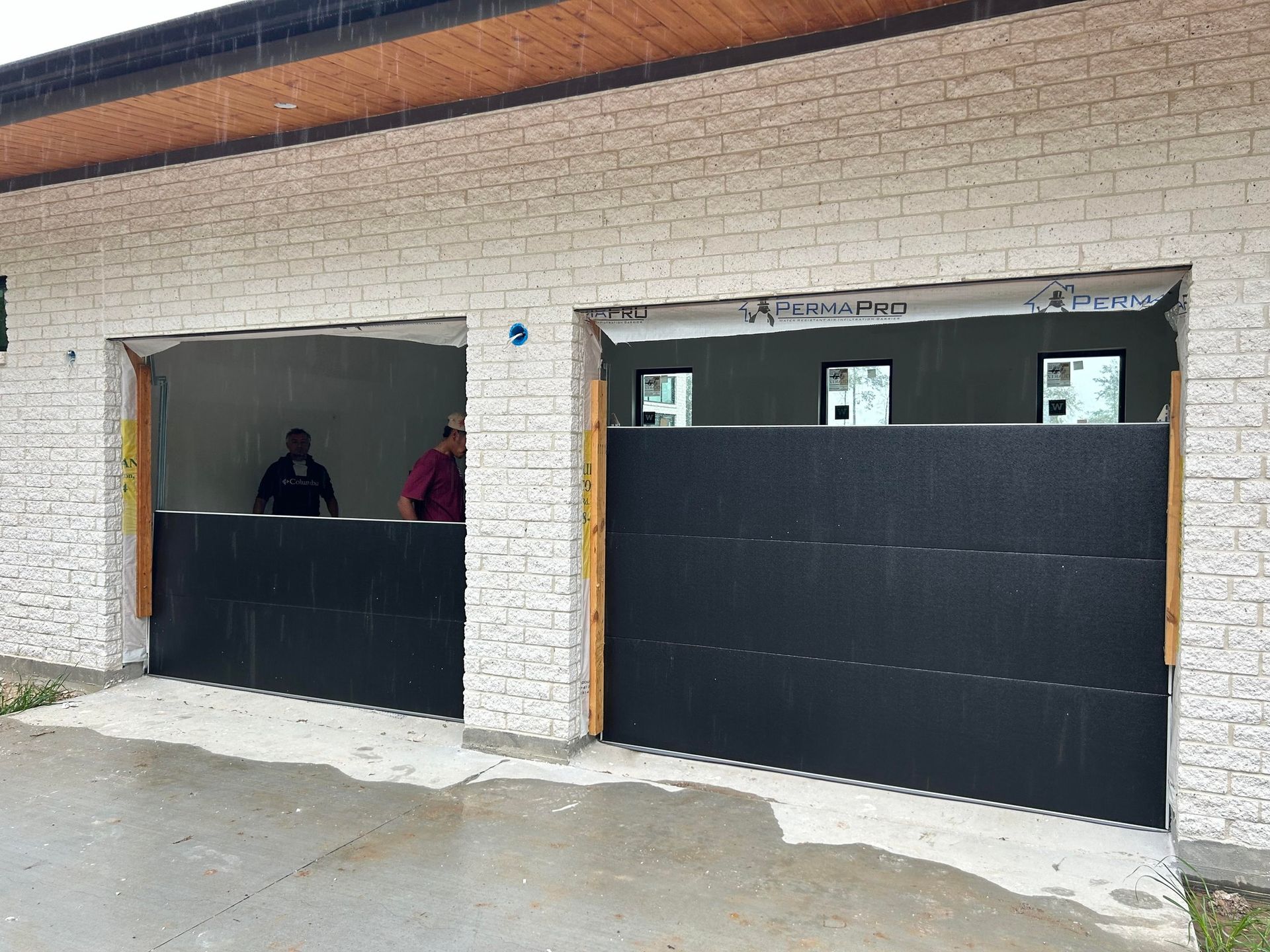 Two garage bays being fitted with new, black insulated door panels under a beige brick building exterior.