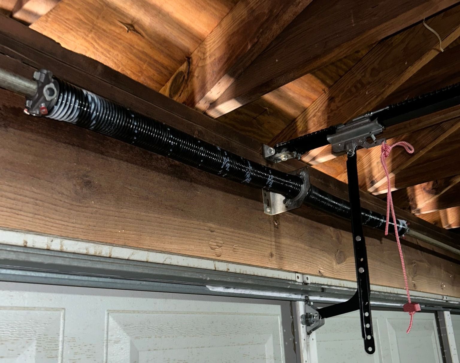A black torsion spring mounted on a wooden beam above a garage door, with a metal mounting bracket and red pull cord.