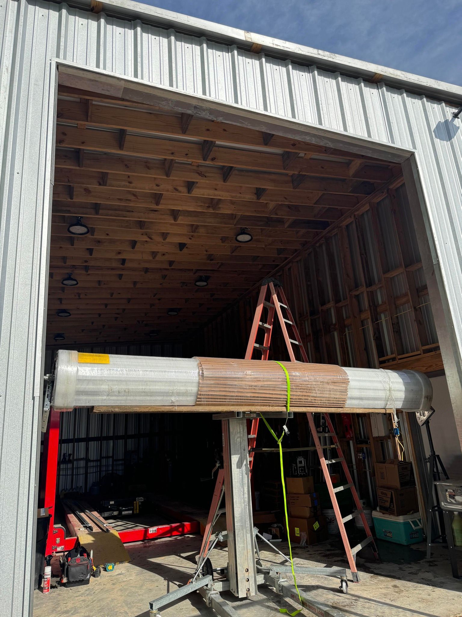 A large, cylindrical object supported by a hoist and straps in the doorway of a metal building with wooden framing.