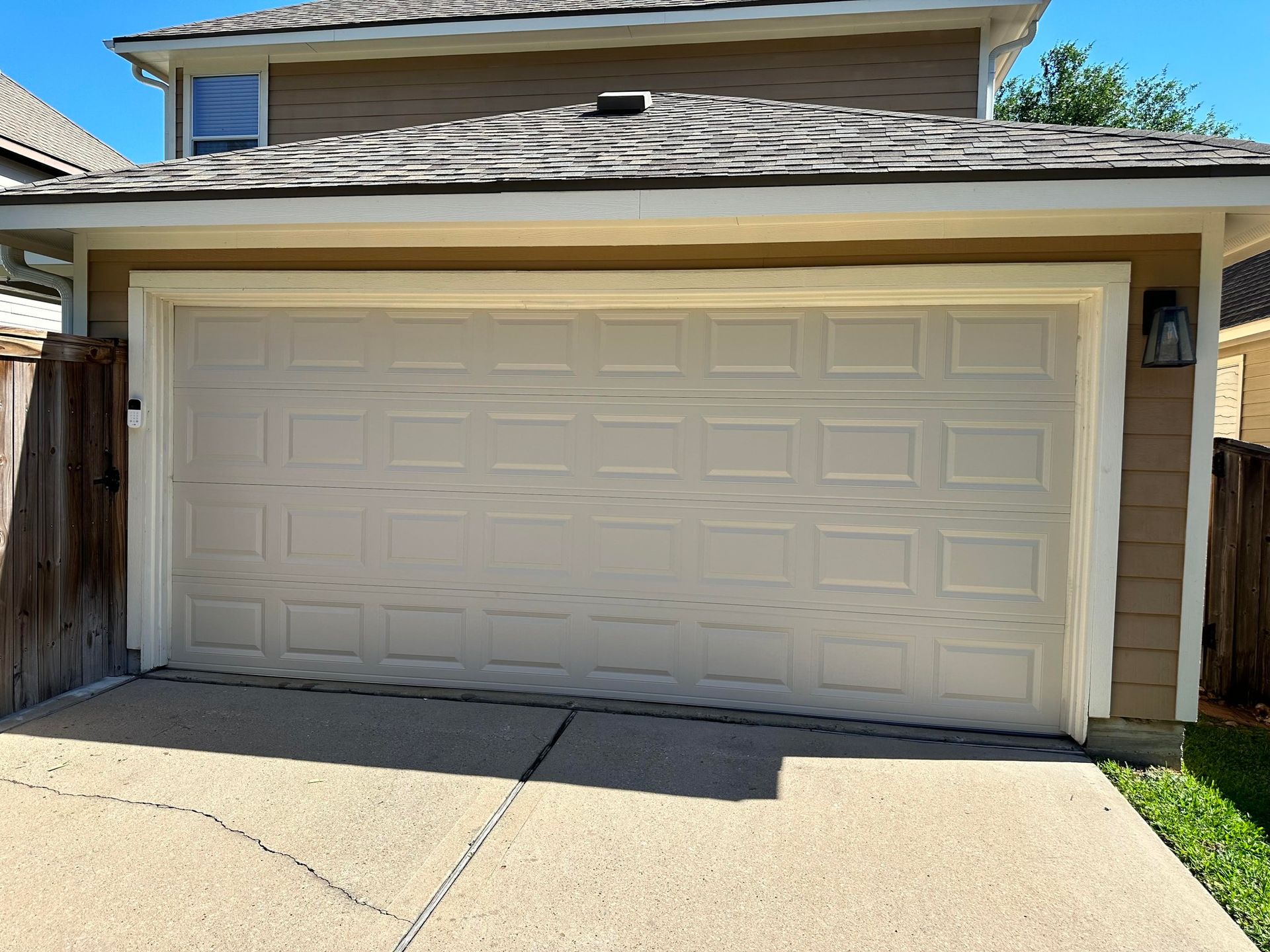 A beige, raised-panel sectional garage door faces a concrete driveway in front of a residential house.