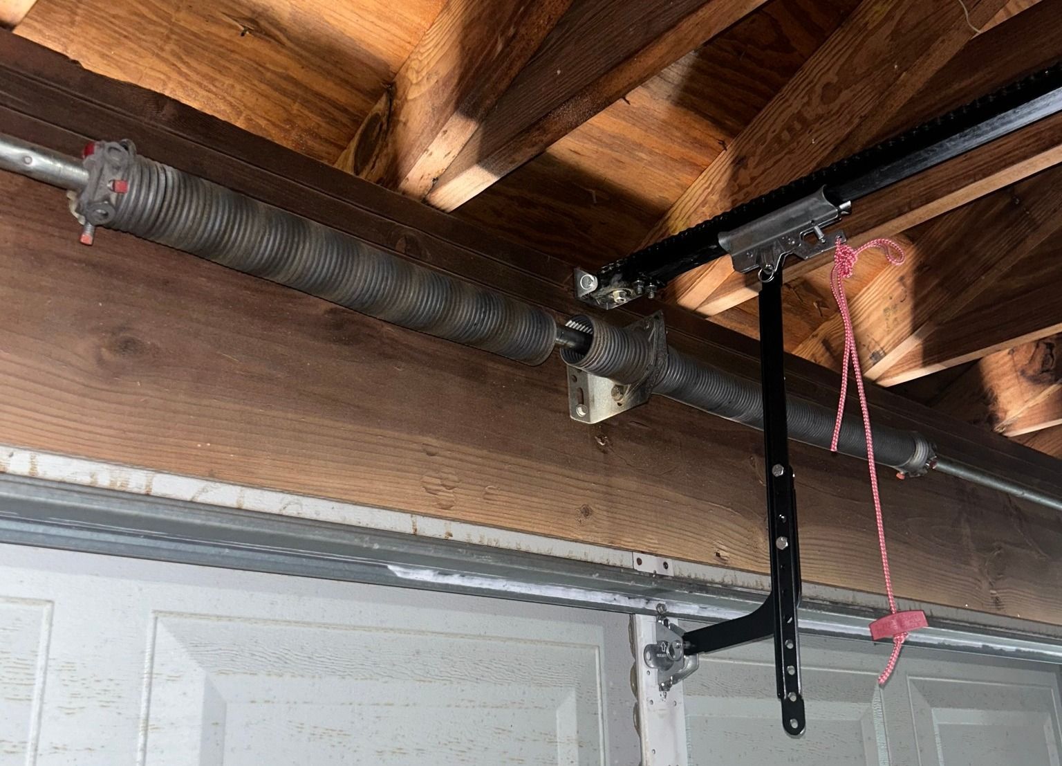 A garage door torsion spring and metal bracket mounted above a door, with a red emergency release cord hanging down.