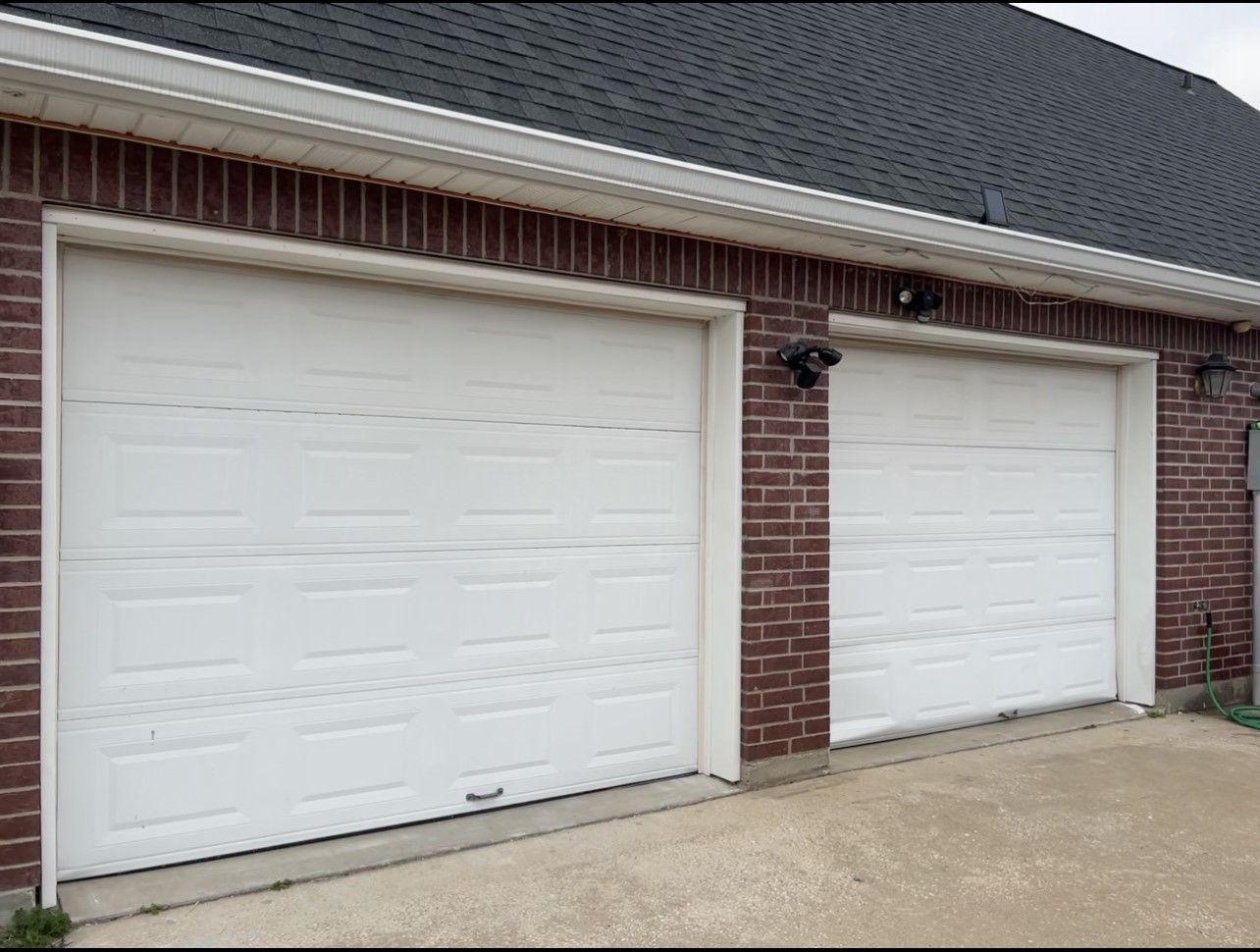 Two white, paneled garage doors set into a dark red brick building with a dark shingled roof.