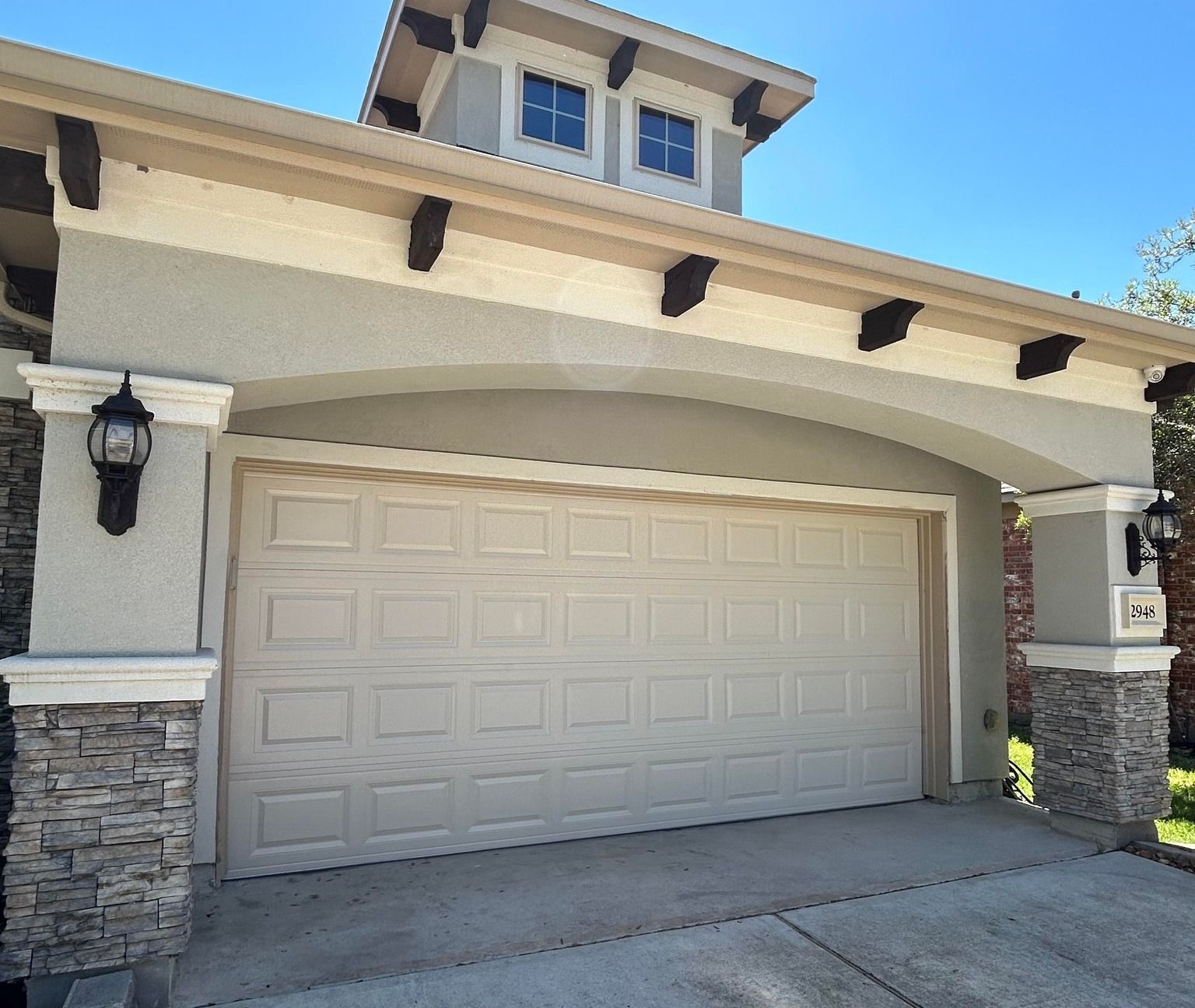 A beige, two-car garage with white trim and stone accents on the pillars, featuring an arched roofline under a blue sky.