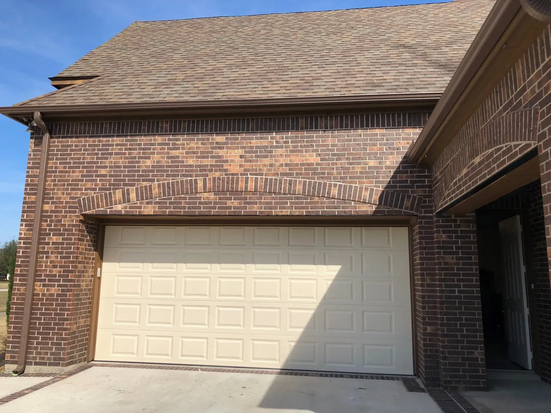 A cream-colored garage door set into a brown brick wall under a shingled roof.