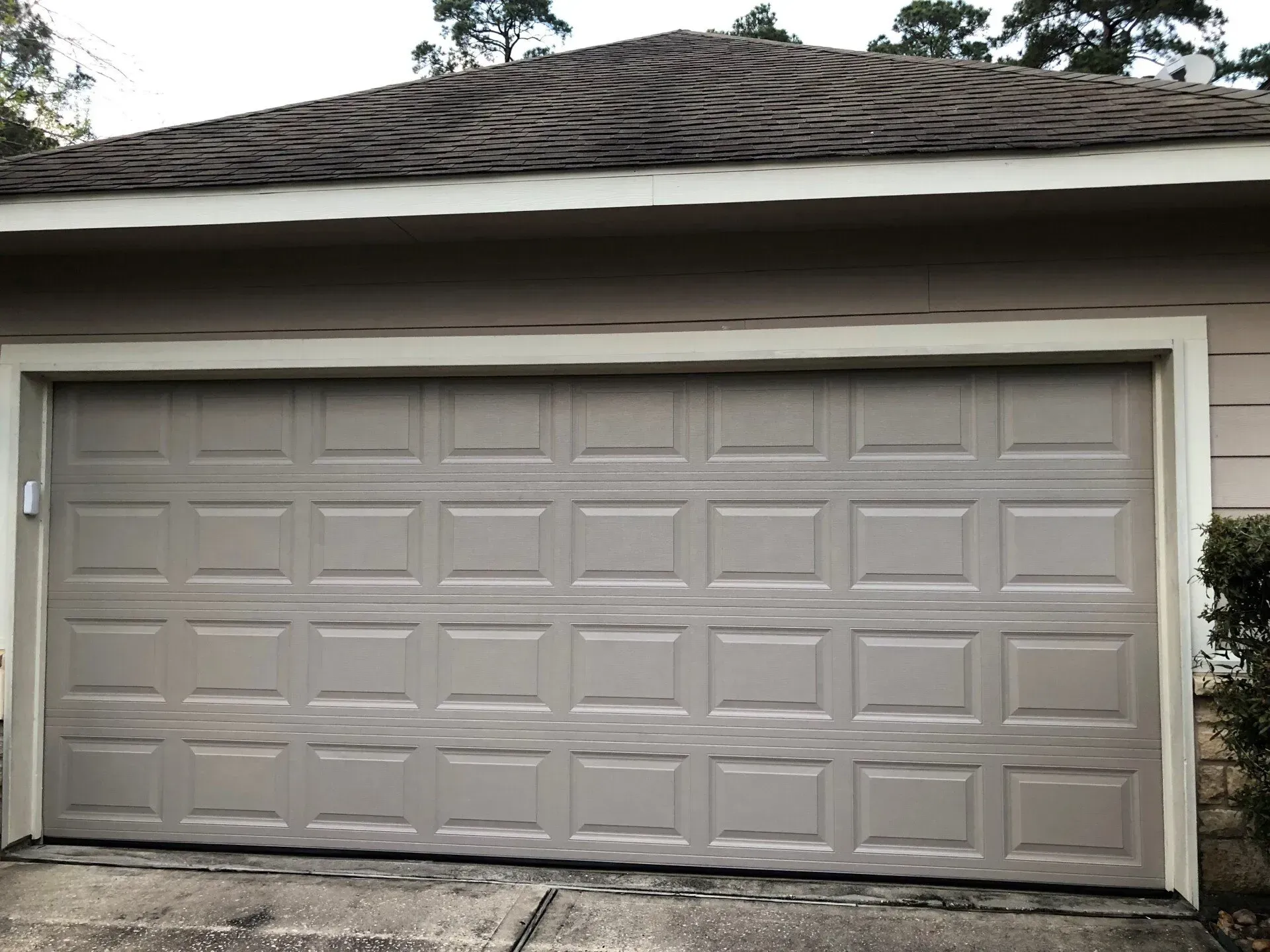 A tan, multi-paneled garage door with a brown shingled roof above, set in a neutral-colored house exterior.