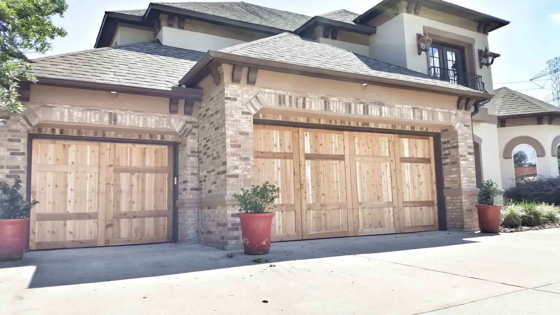 A three-car garage with natural wood doors set in a stone and stucco home under a blue sky, flanked by red planters.