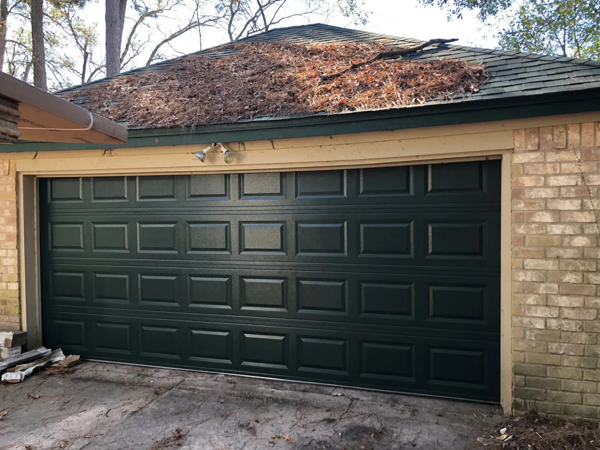 A dark green garage door on a brick building with a roof covered in fallen brown leaves.