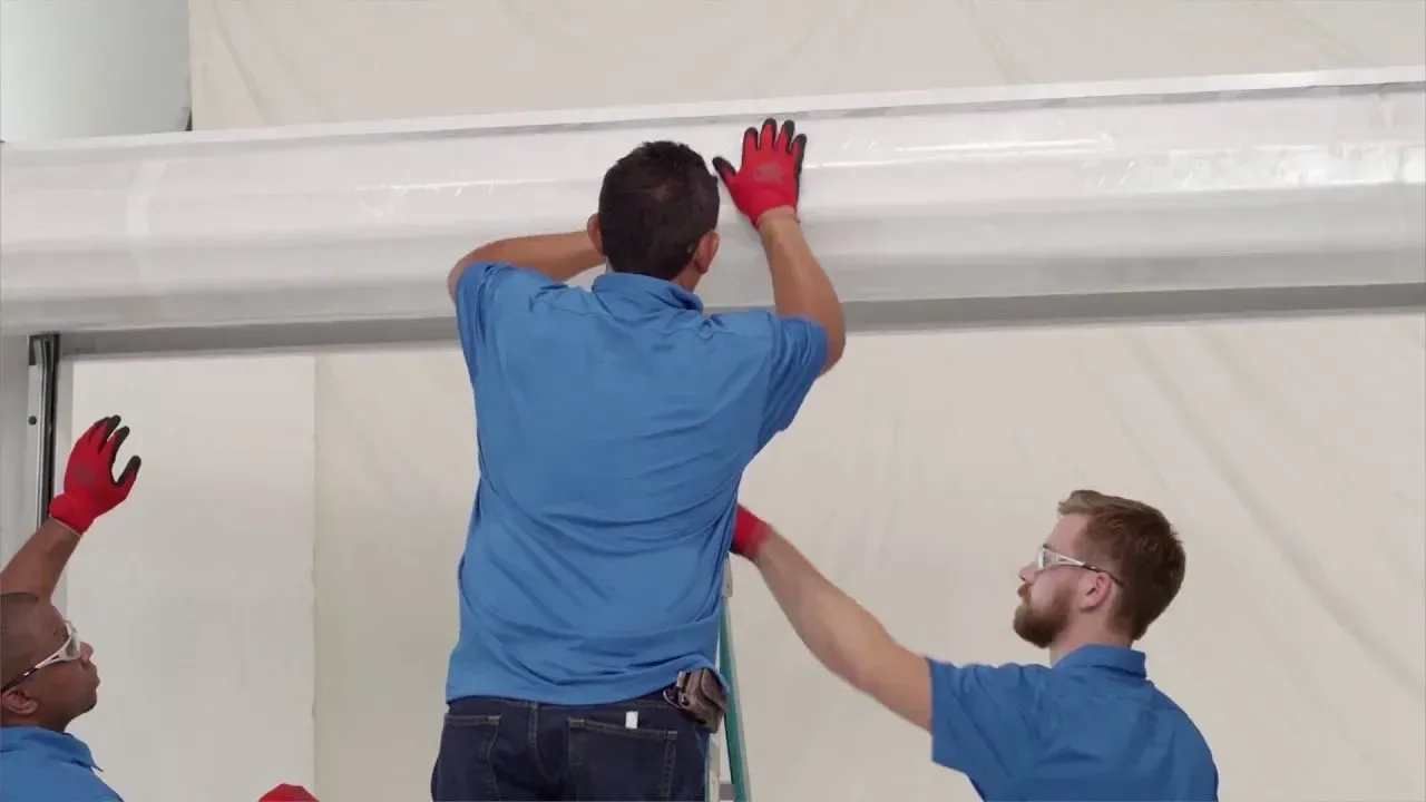 Three technicians in blue shirts and red gloves work together to install a large white cylindrical component.