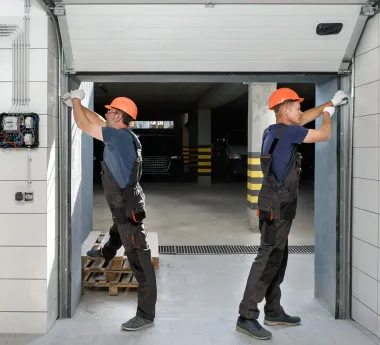 Two workers in hard hats and workwear install a garage door in an industrial facility.