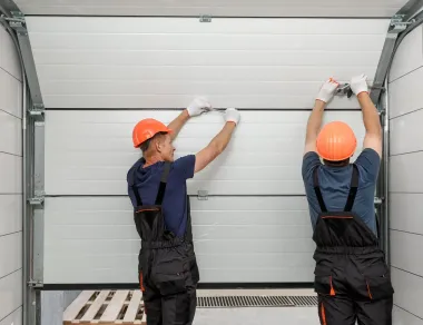 Two technicians in hard hats and work coveralls installing a sectional garage door.