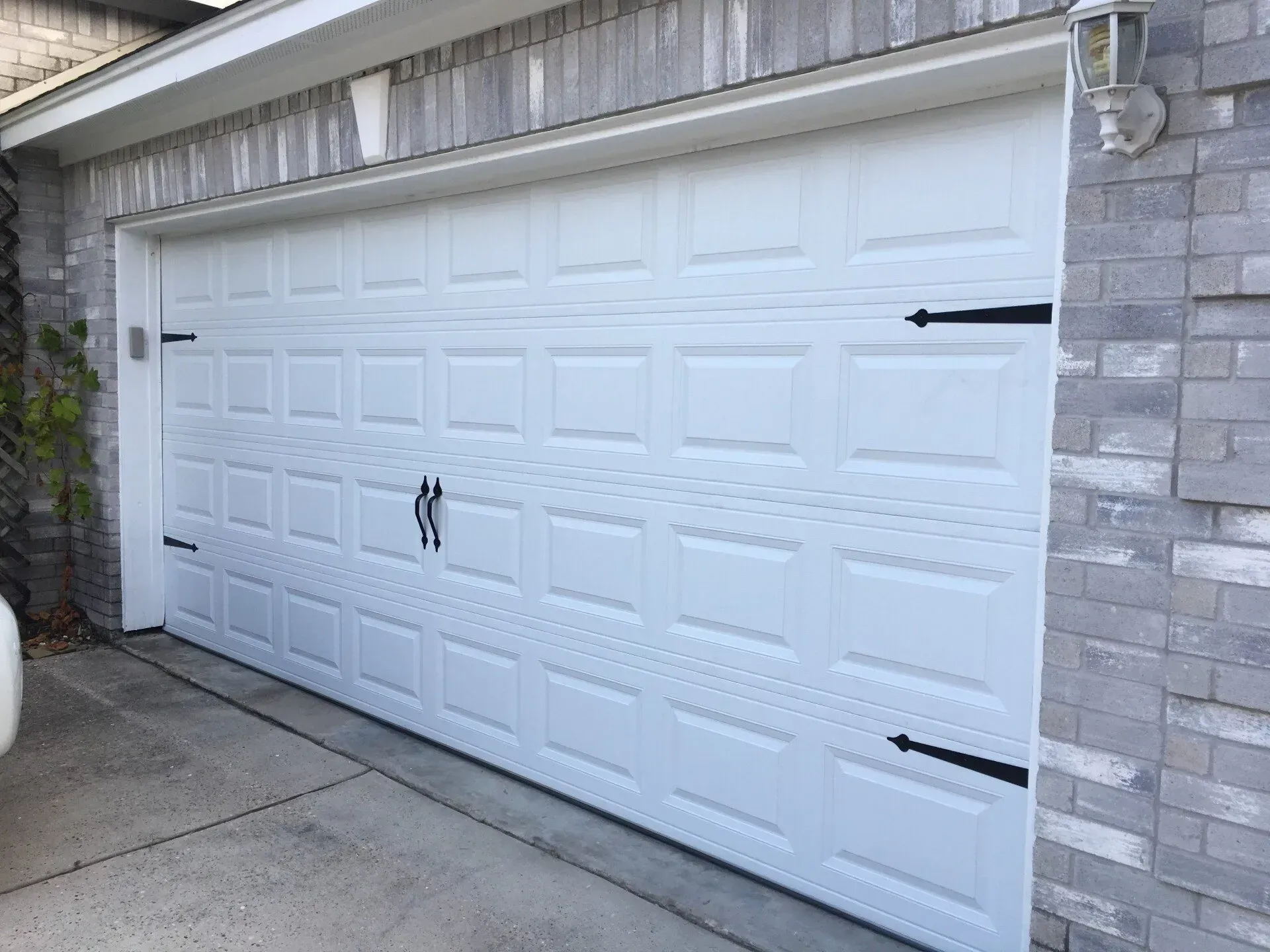 A white garage door with black decorative hinges and handles, installed on a house with a gray brick exterior.