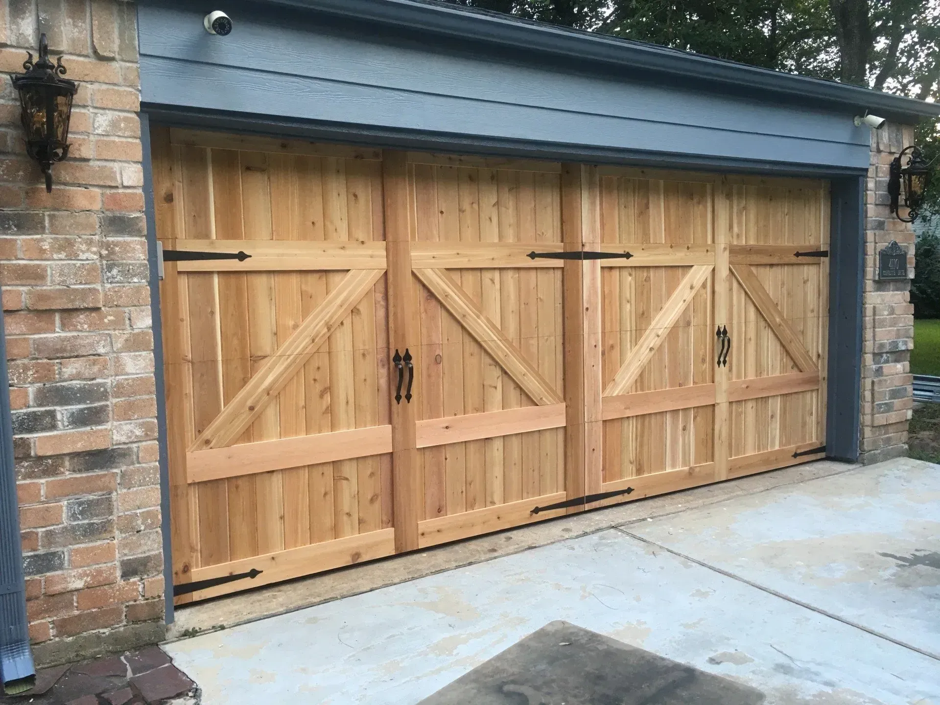 Wooden carriage-style garage door with black decorative hinges and handles, installed in a brick house exterior.