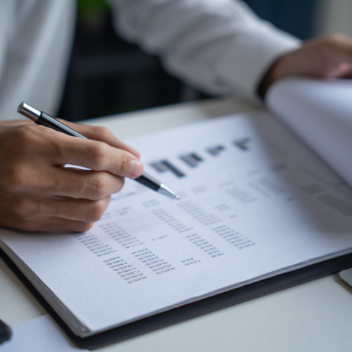 Person's hands holding a pen, examining a document with data charts and text.