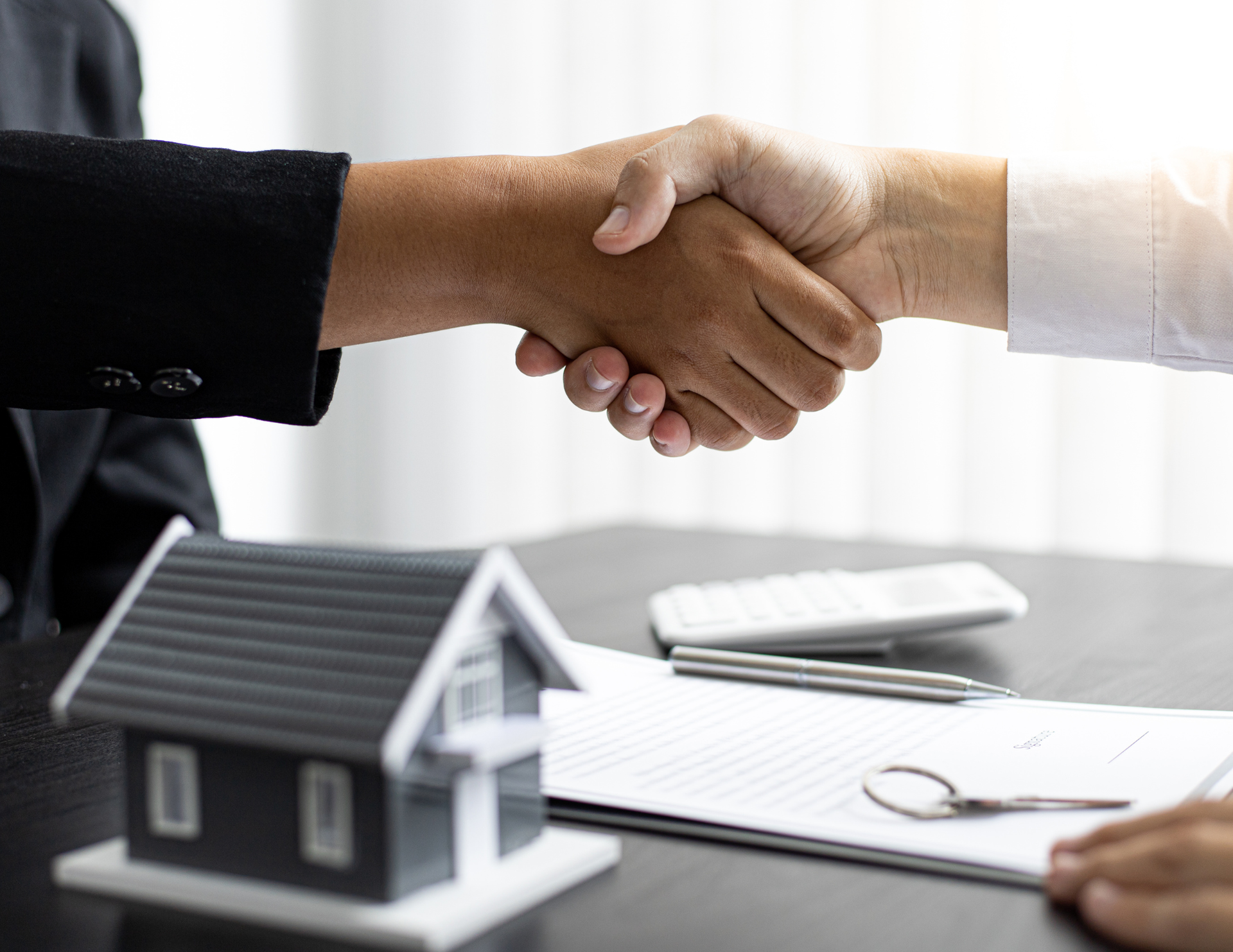 Handshake seals real estate deal with house model, documents, and calculator on the desk.