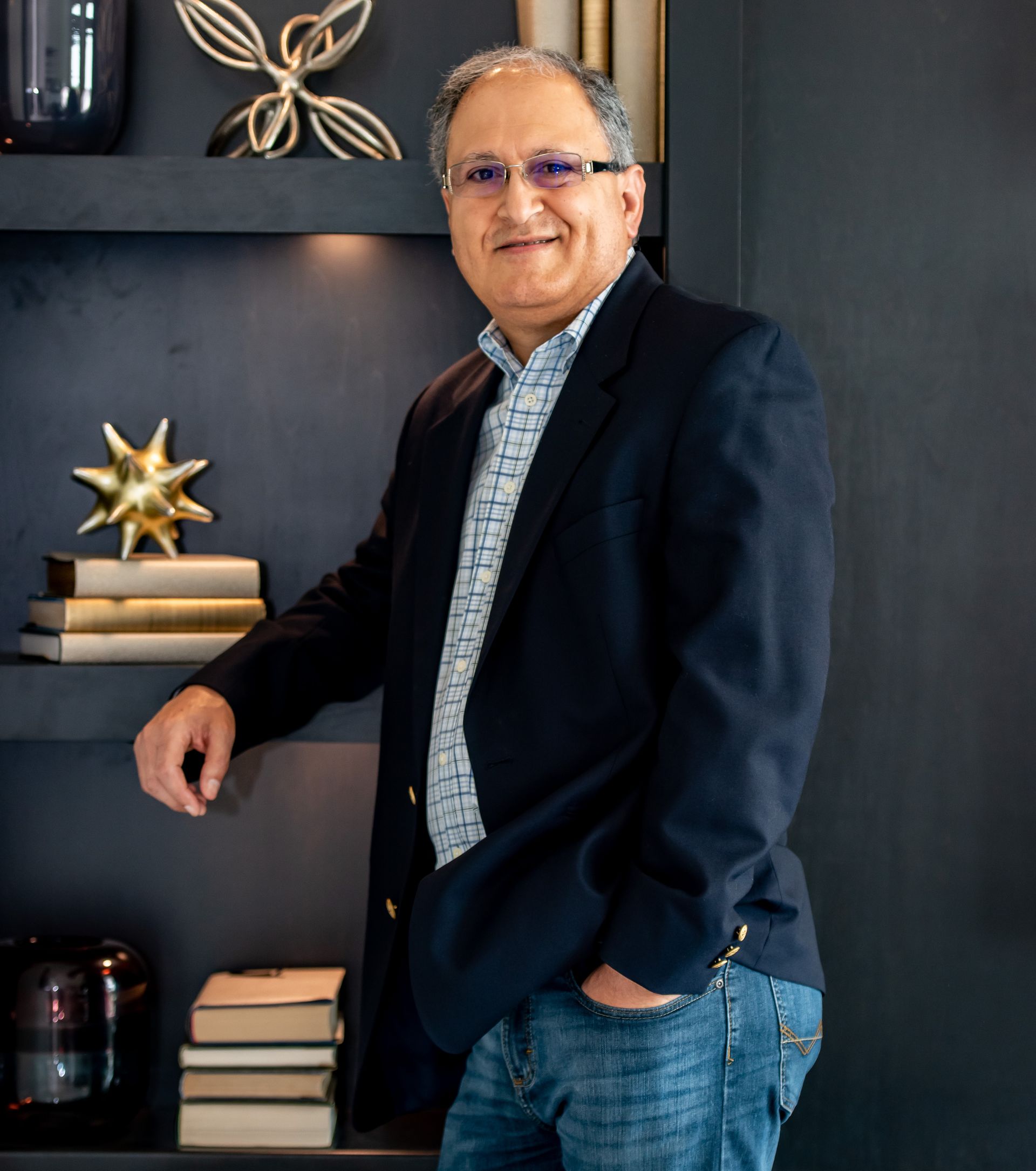 Man in blazer and jeans, leaning on a shelf, smiling. Dark wall with art objects.