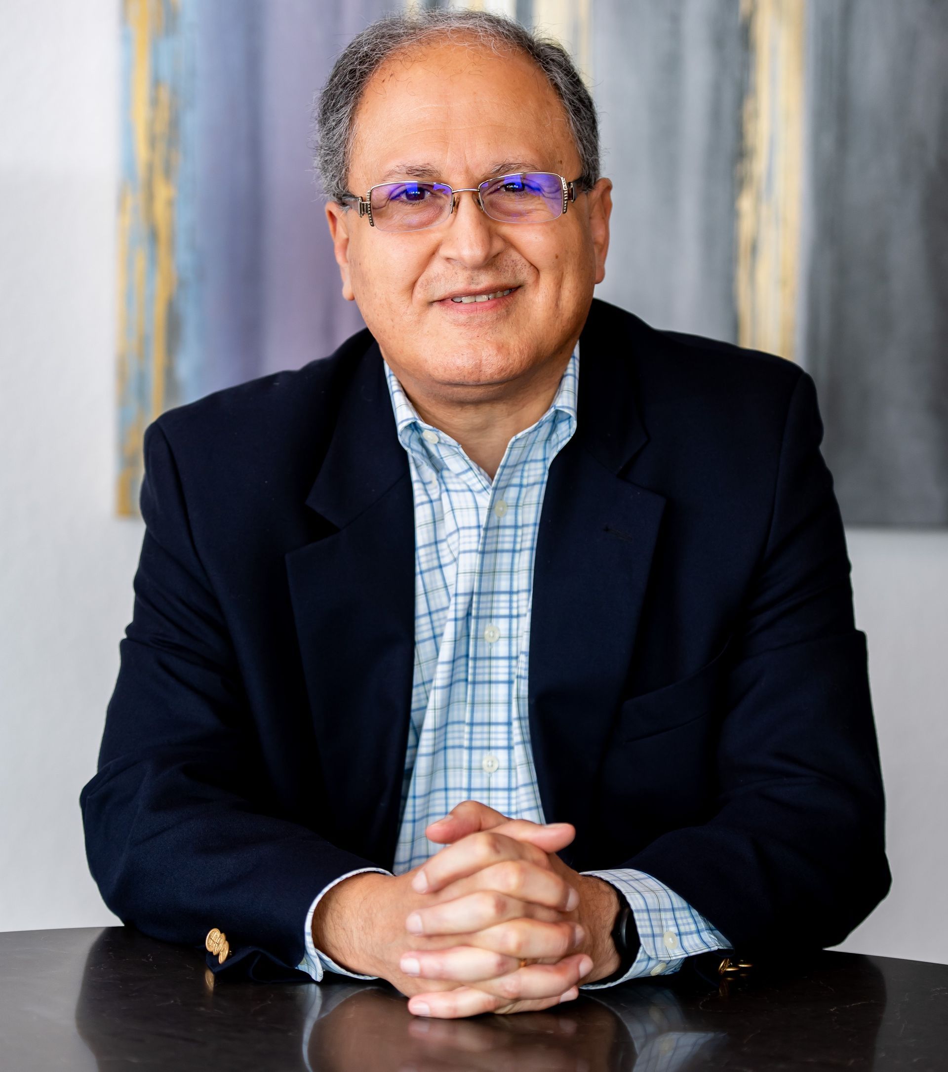 Man with glasses, navy blazer, and blue plaid shirt, seated with hands clasped, smiling at camera.