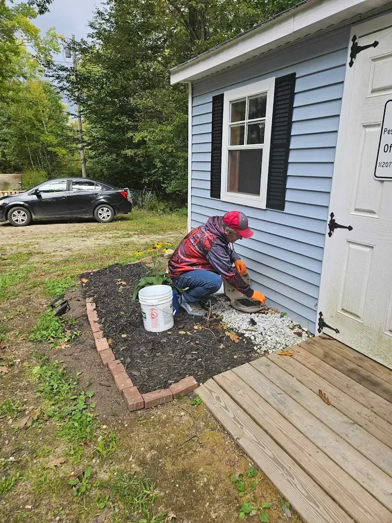 Person painting the house’s exterior siding near a doorway, with a bucket and car visible in the yard.
