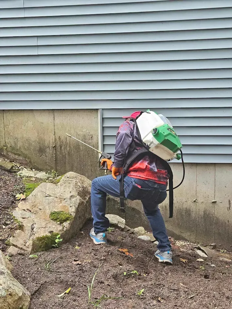 Worker kneeling with backpack sprayer beside house foundation, applying treatment near rocks and dirt