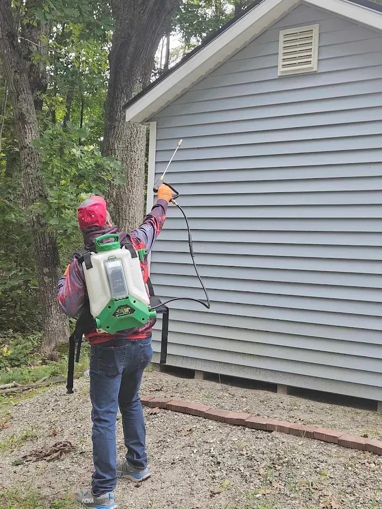 Person using a backpack sprayer to treat the side of a gray house outdoors