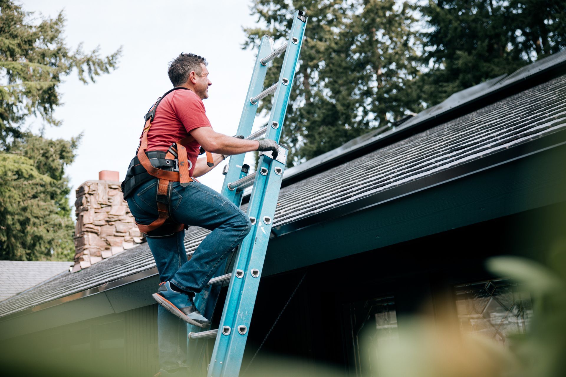 A worker in a red shirt climbs a blue ladder to reach the roof of a house.