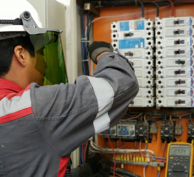 Technician inspecting an electrical panel with wiring and circuit breakers, wearing a hard hat and face shield