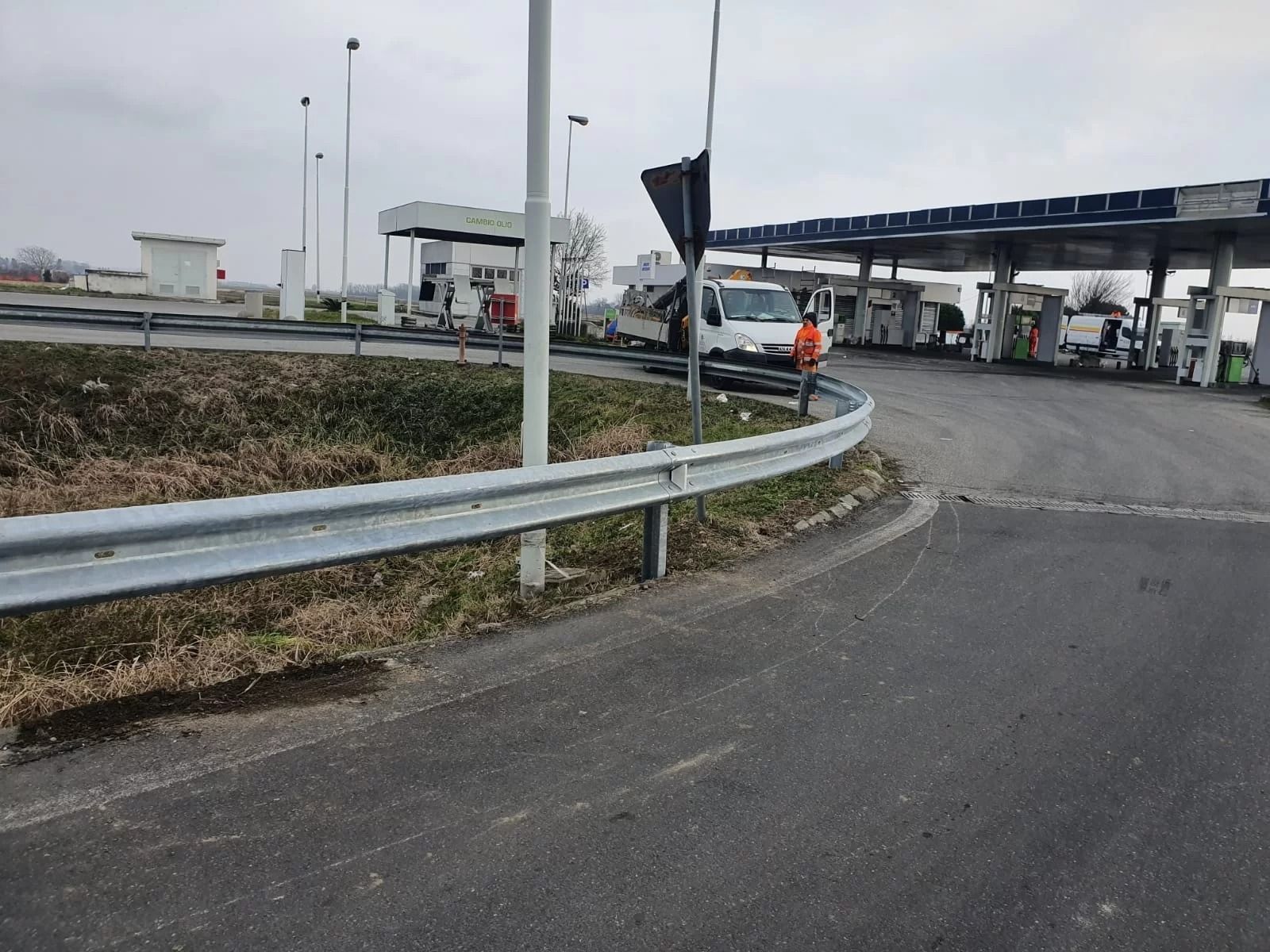 Un operaio con un giubbotto arancione è in piedi vicino a un furgone bianco presso una stazione di servizio lungo la strada, con una barriera metallica di protezione in primo piano.