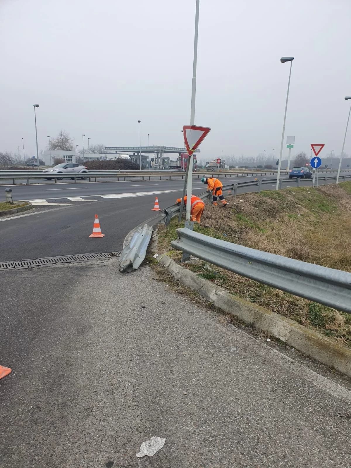 Due operai con indumenti di sicurezza arancioni brillanti riparano un guardrail danneggiato a bordo strada vicino a un incrocio.