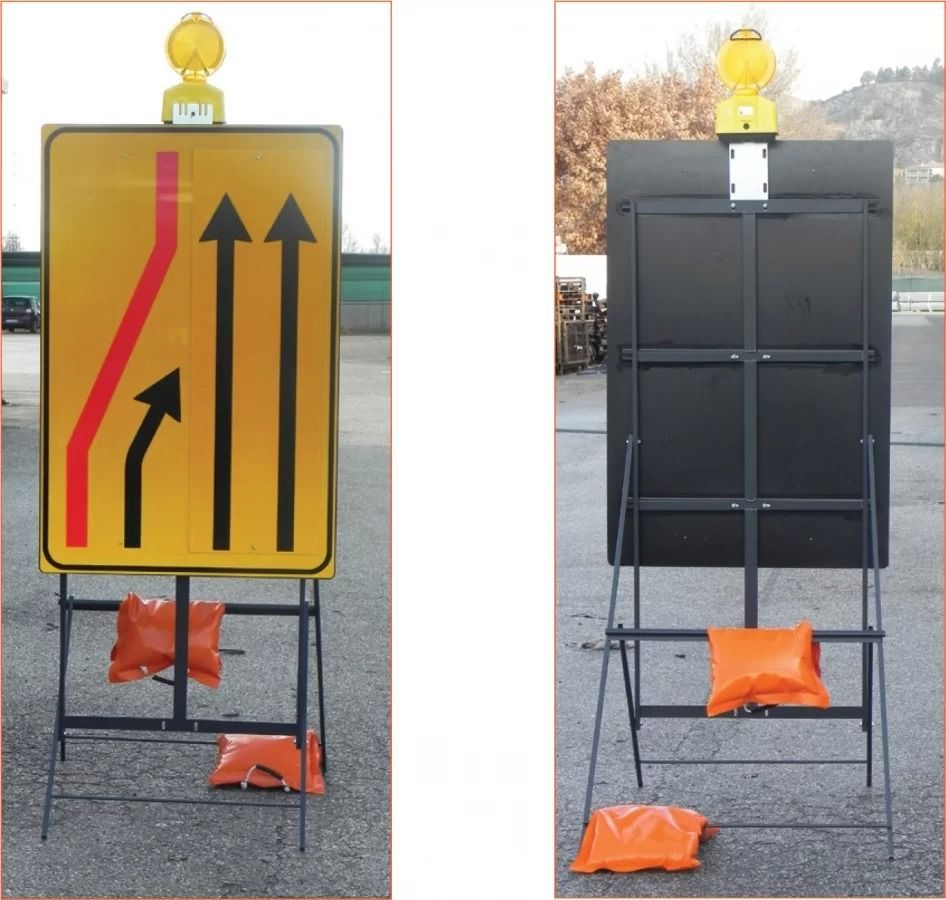 Un cartello giallo di lavori stradali che indica tre corsie, con la corsia più a sinistra che si immette a destra, montato su un supporto con sacchi di sabbia.