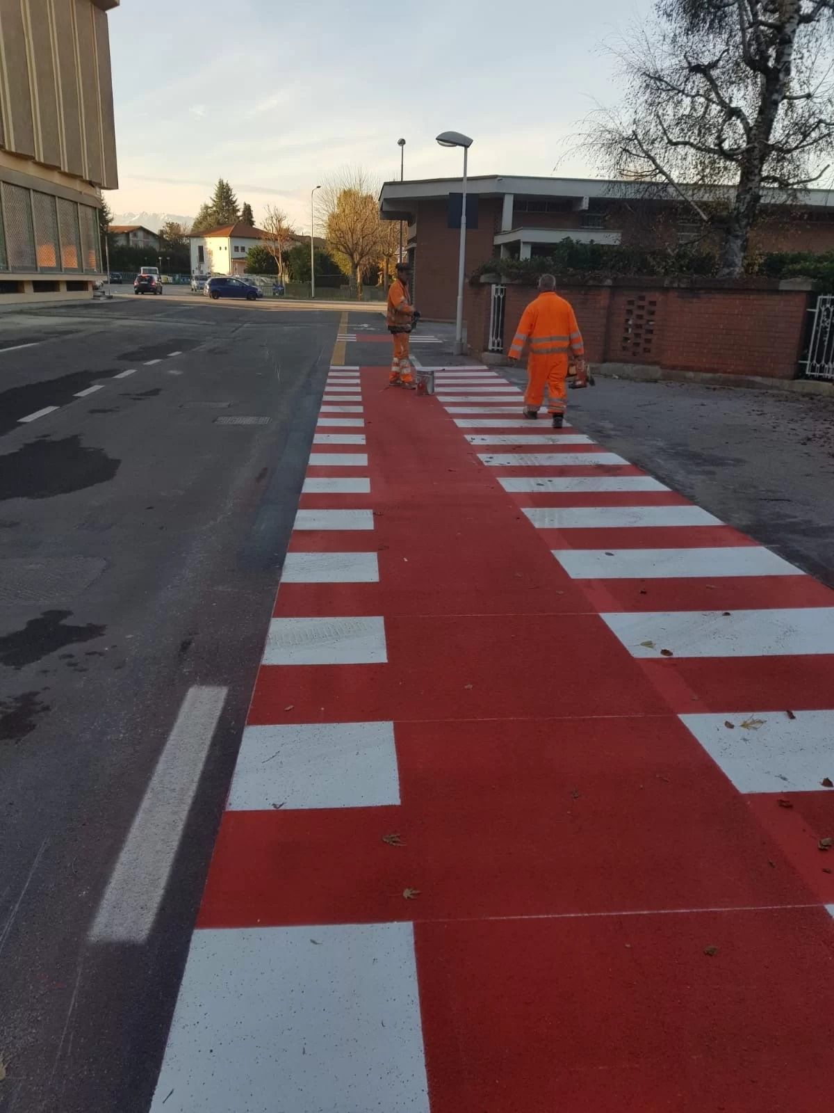 Due operai con indumenti arancioni ad alta visibilità dipingono un attraversamento pedonale a strisce rosse e bianche su una strada asfaltata.