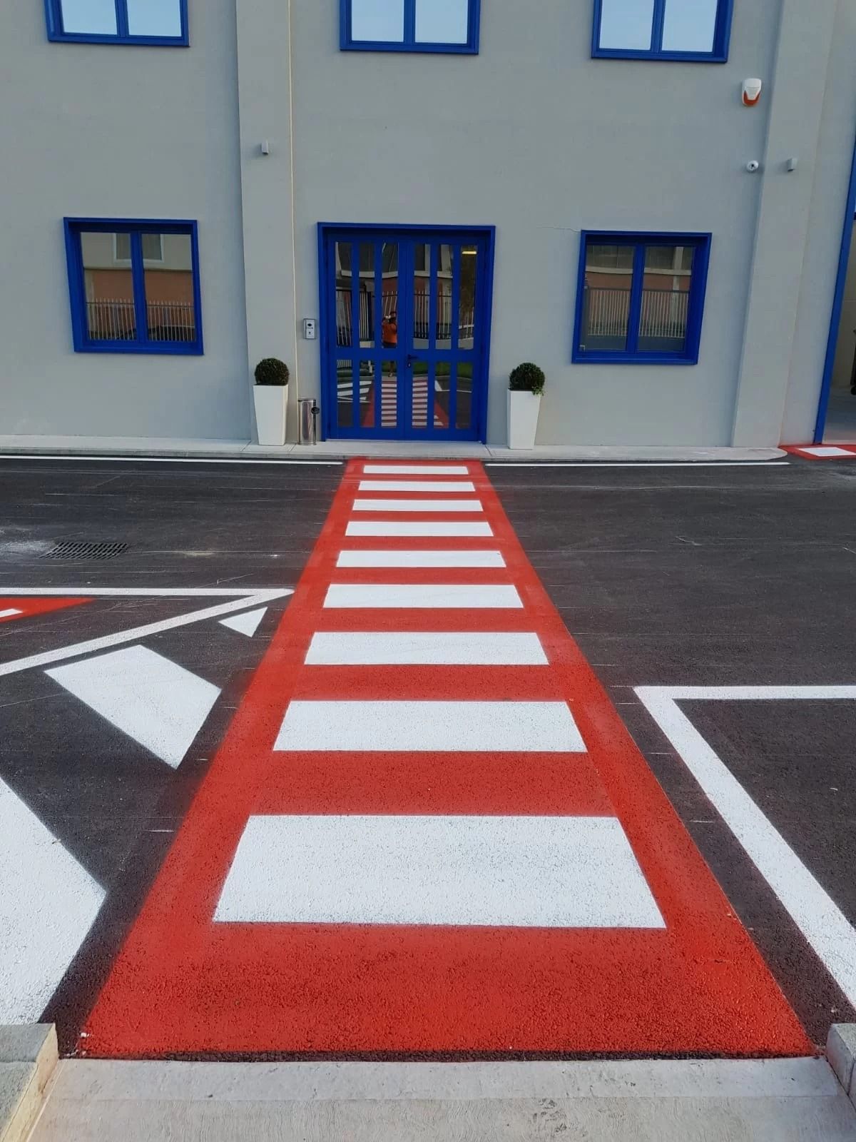 Un attraversamento pedonale rosso acceso con strisce bianche conduce all'ingresso di un edificio grigio con finestre incorniciate di blu.