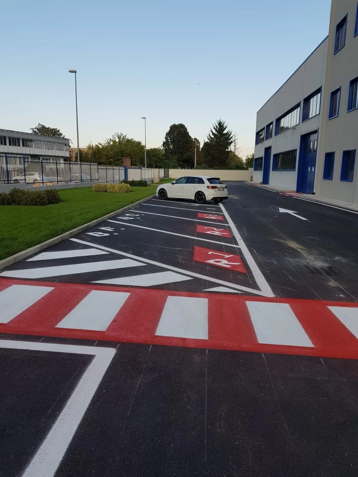 Un parcheggio con attraversamenti pedonali a strisce rosse e bianche e posti auto riservati, con un'auto bianca sullo sfondo.