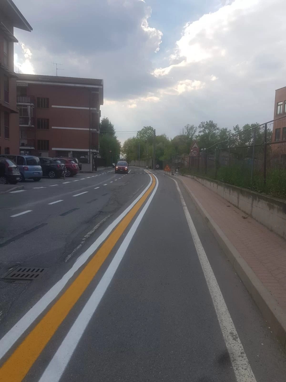 Una veduta stradale con una pista ciclabile dedicata sulla destra, segnalata da linee bianche e gialle, accanto a edifici e auto parcheggiate.