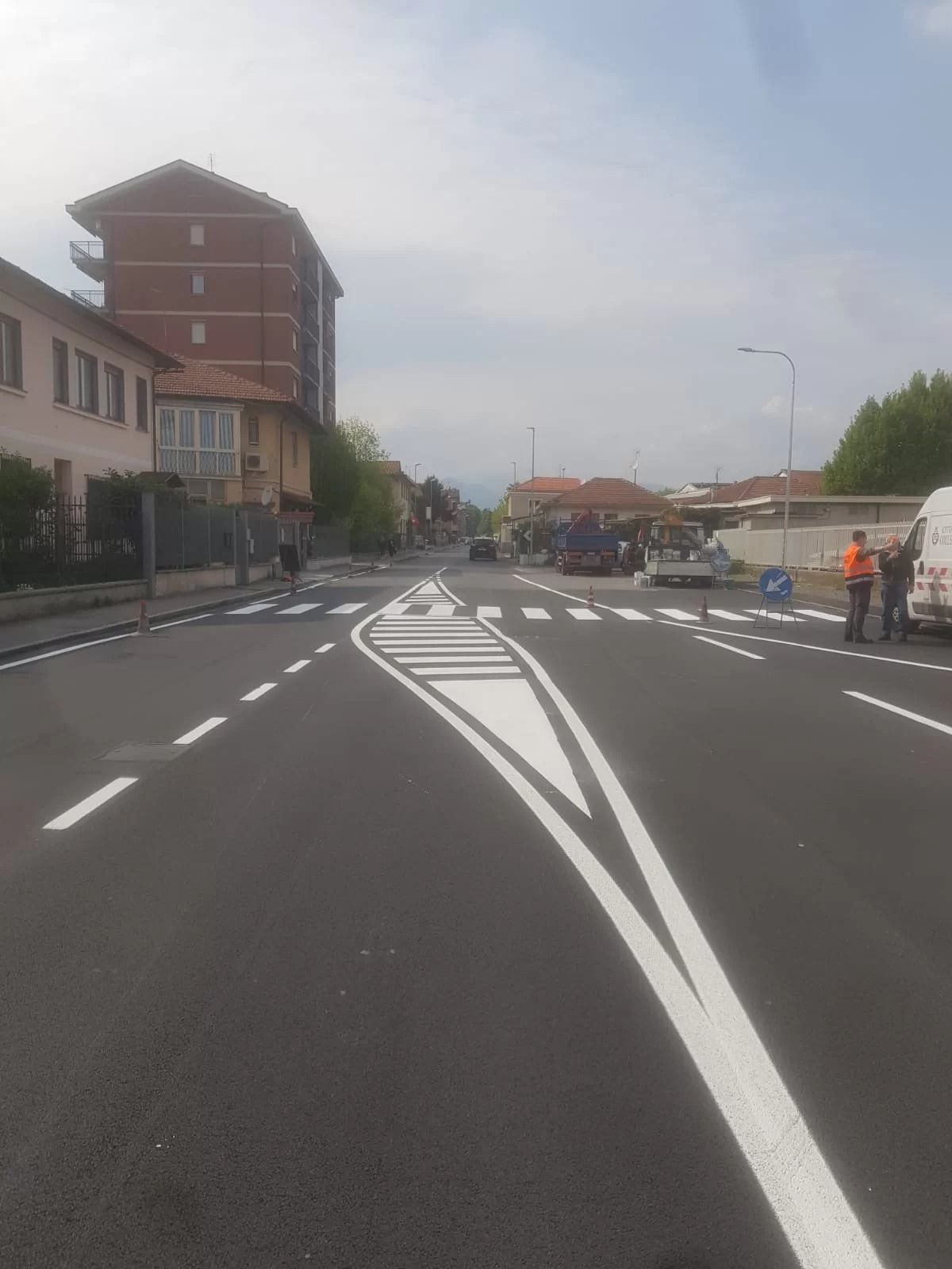 Una strada appena asfaltata con segnaletica orizzontale bianca e un'isola pedonale centrale, con edifici e operai ai lati.