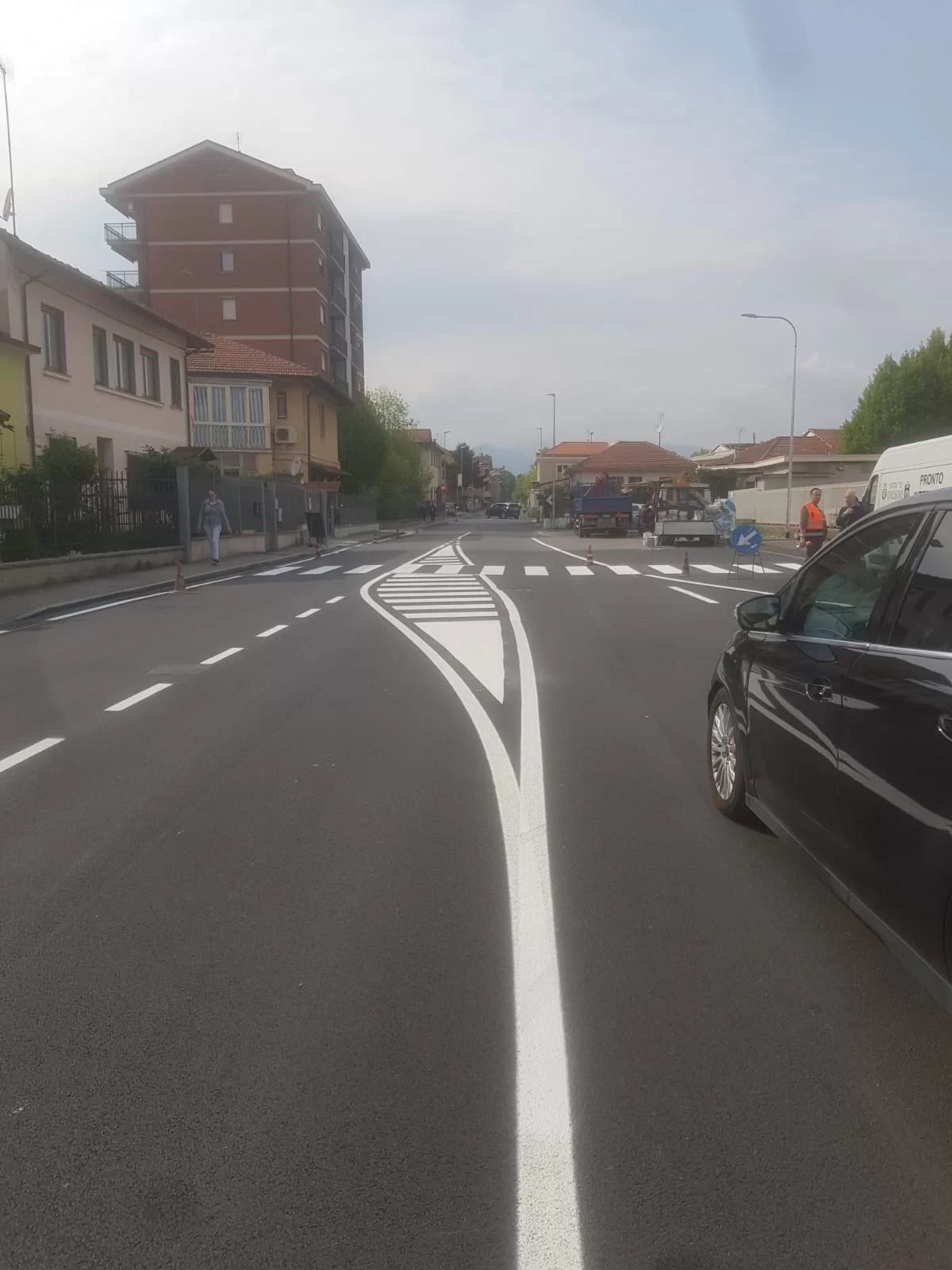 Vista di una strada asfaltata con un'isola spartitraffico bianca al centro, che conduce a un attraversamento pedonale segnalato.