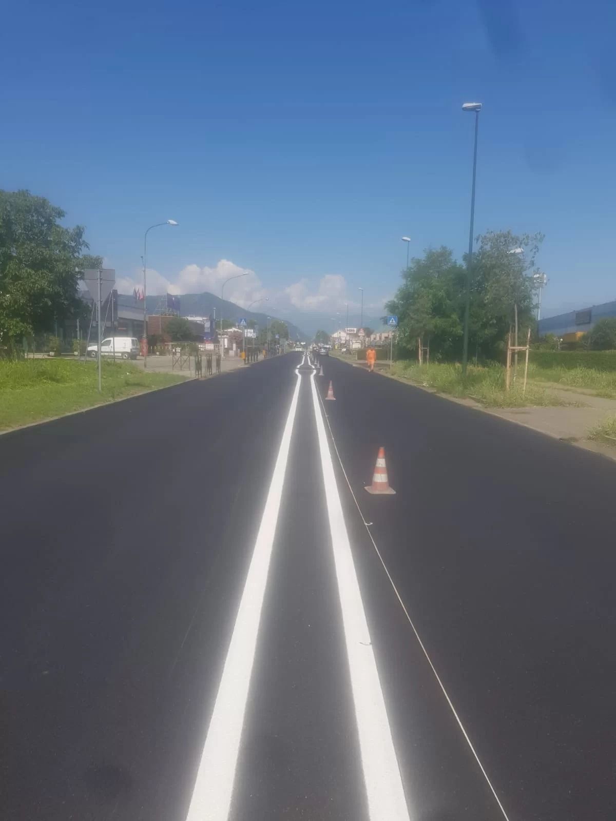 Una strada dritta, appena asfaltata, con doppia linea bianca e coni stradali arancioni, sotto un cielo azzurro e limpido.