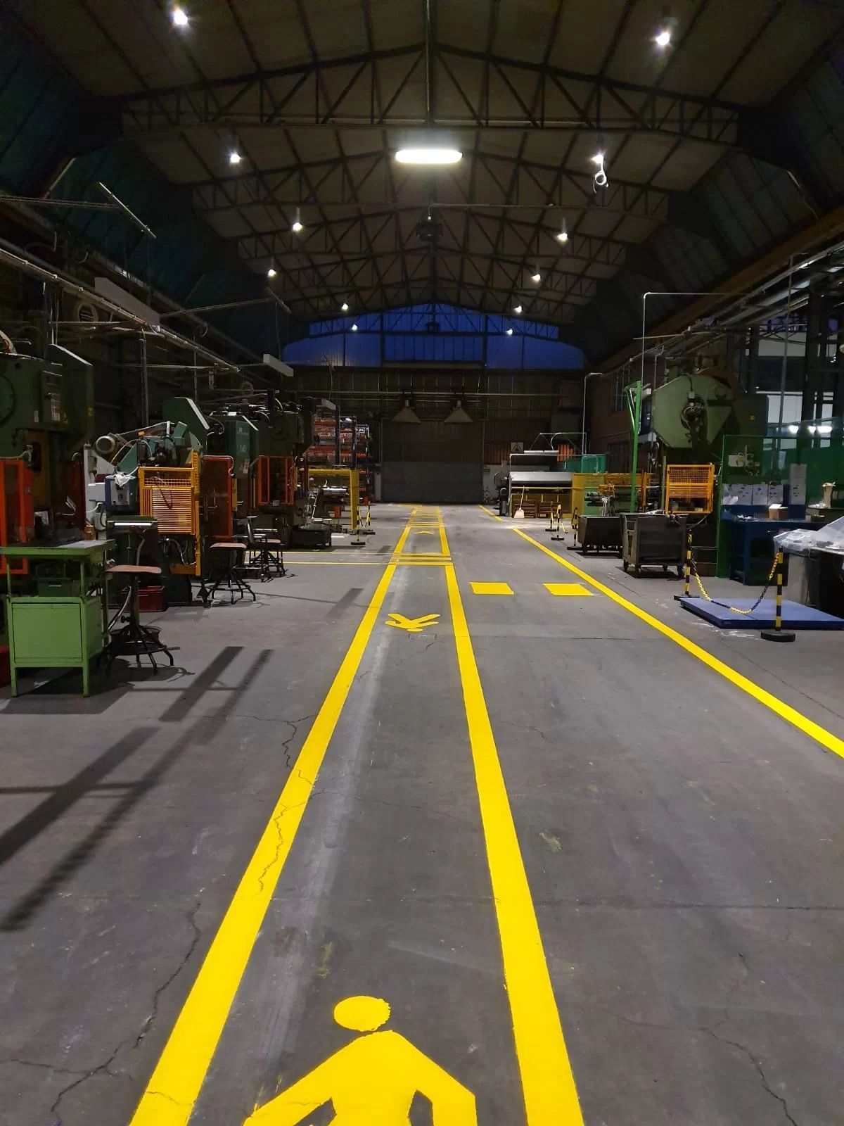 Un percorso pedonale segnalato da linee gialle con il simbolo del pedone su una pavimentazione in cemento all'interno di un grande stabilimento industriale.
