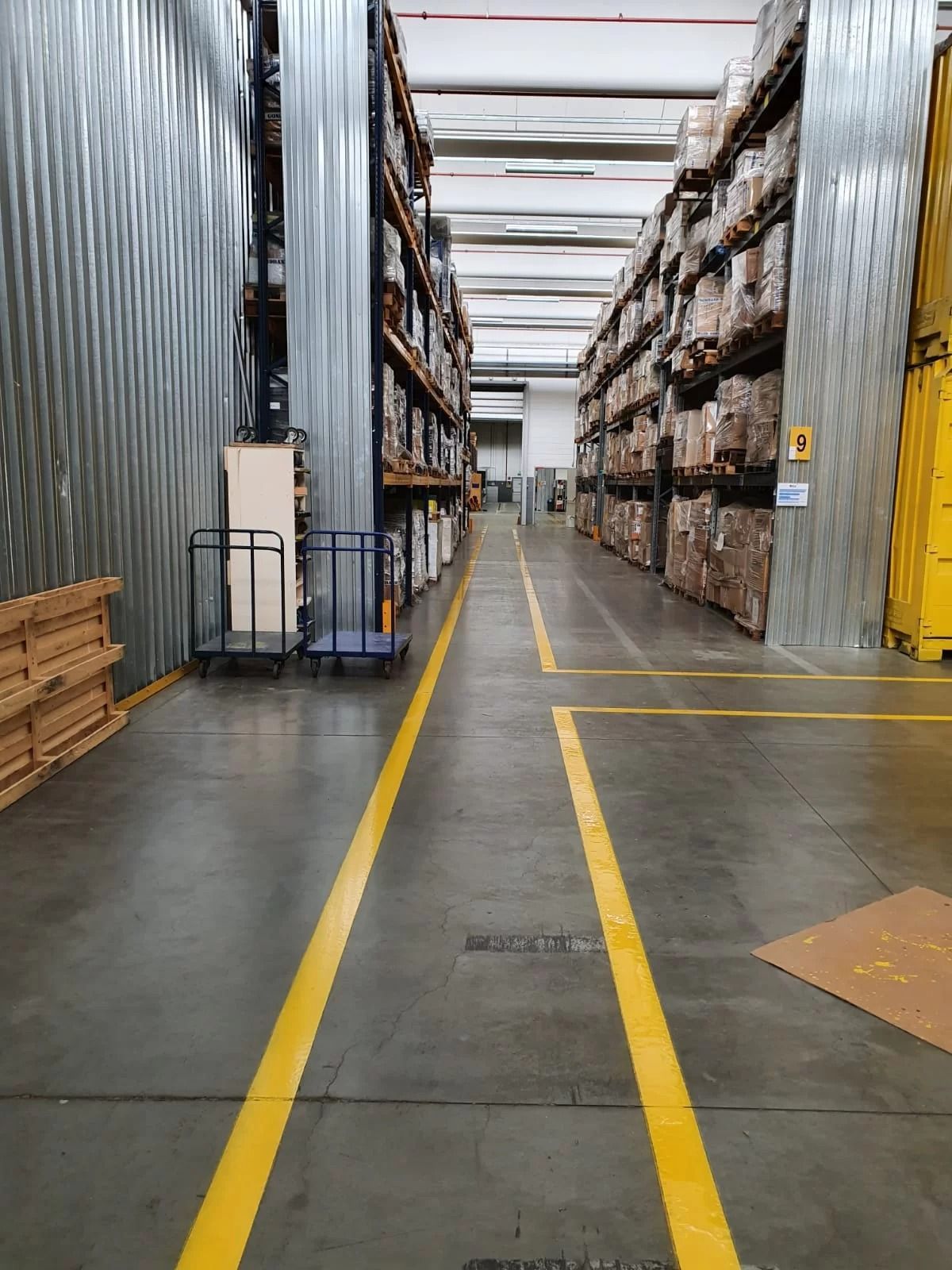 Un corridoio di magazzino con scaffalature alte su entrambi i lati, pareti metalliche e segnaletica di sicurezza gialla sul pavimento.