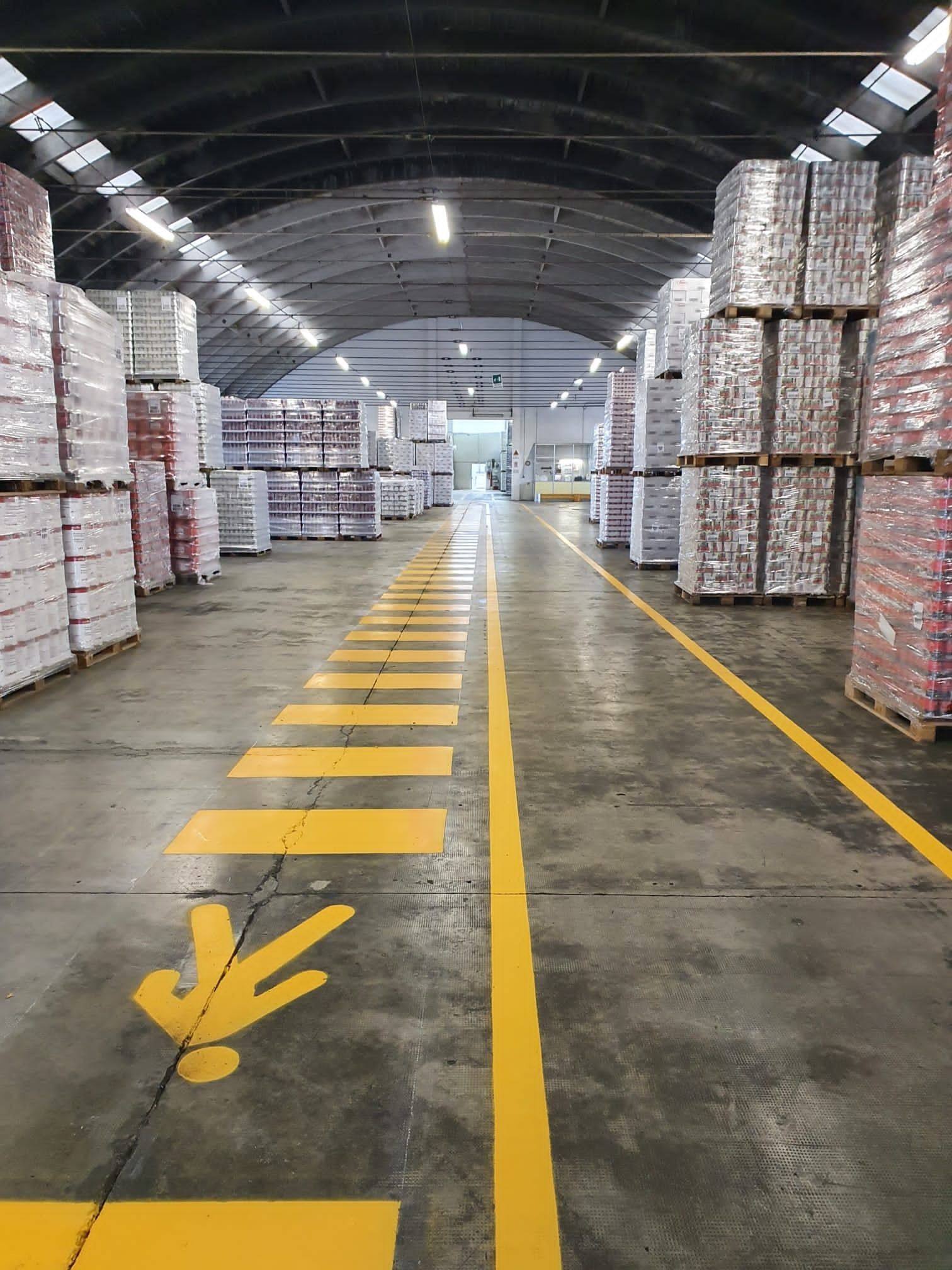 Un corridoio di un magazzino con alte pile di pallet e un percorso pedonale segnalato da una striscia gialla dipinta sul pavimento.