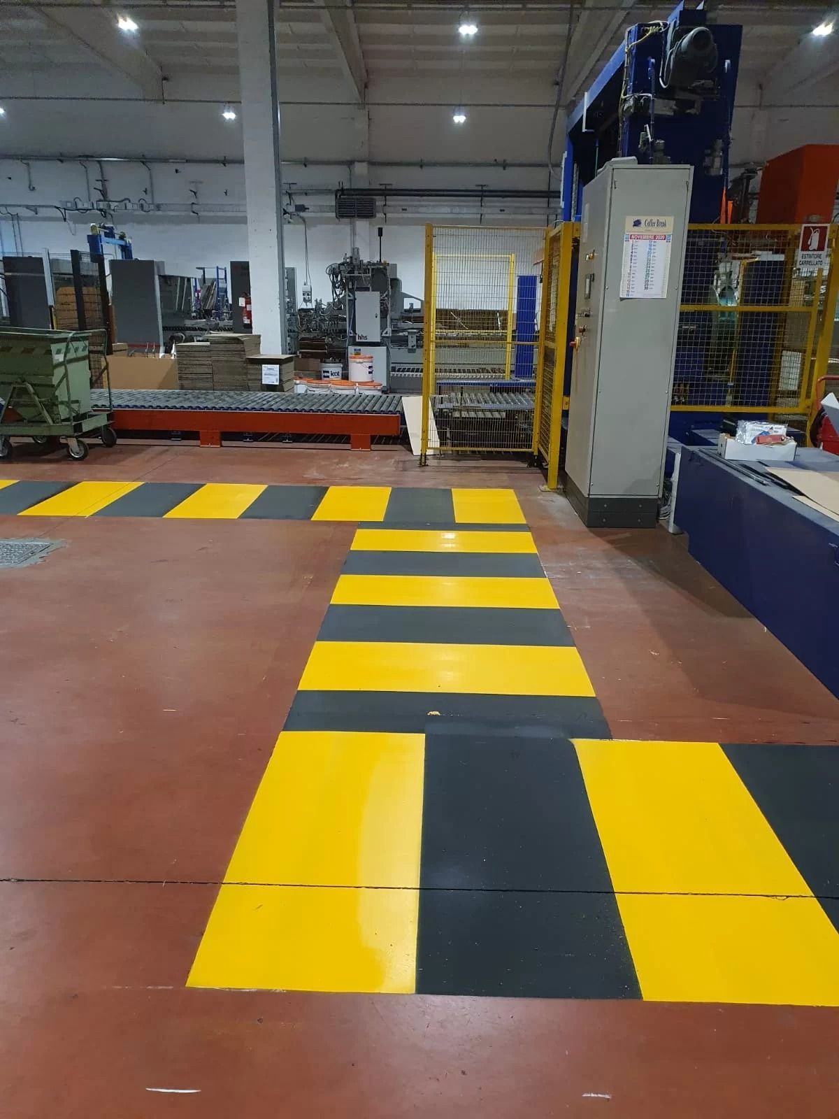 Il pavimento di un magazzino presenta una passerella pedonale a strisce gialle e nere che delimita un percorso intorno ai macchinari industriali.