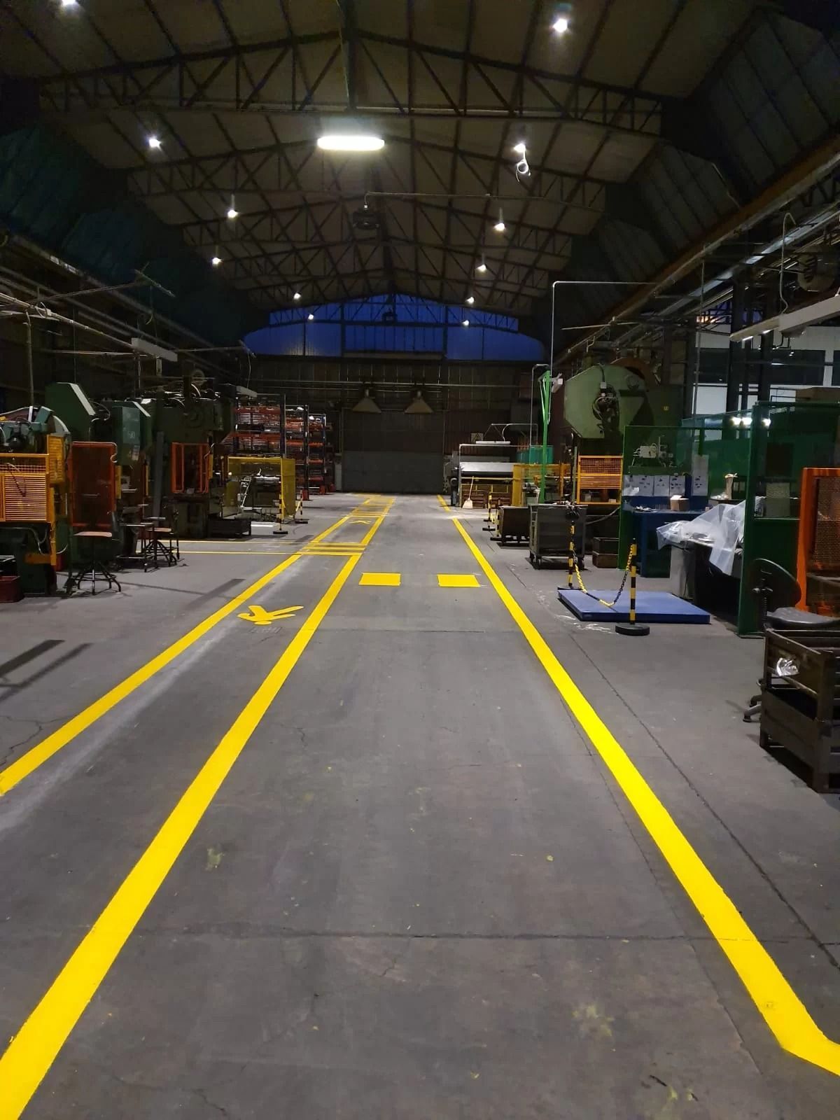Interno di una fabbrica con macchinari che fiancheggiano un corridoio centrale delimitato da linee dipinte di giallo brillante e simboli sul pavimento.