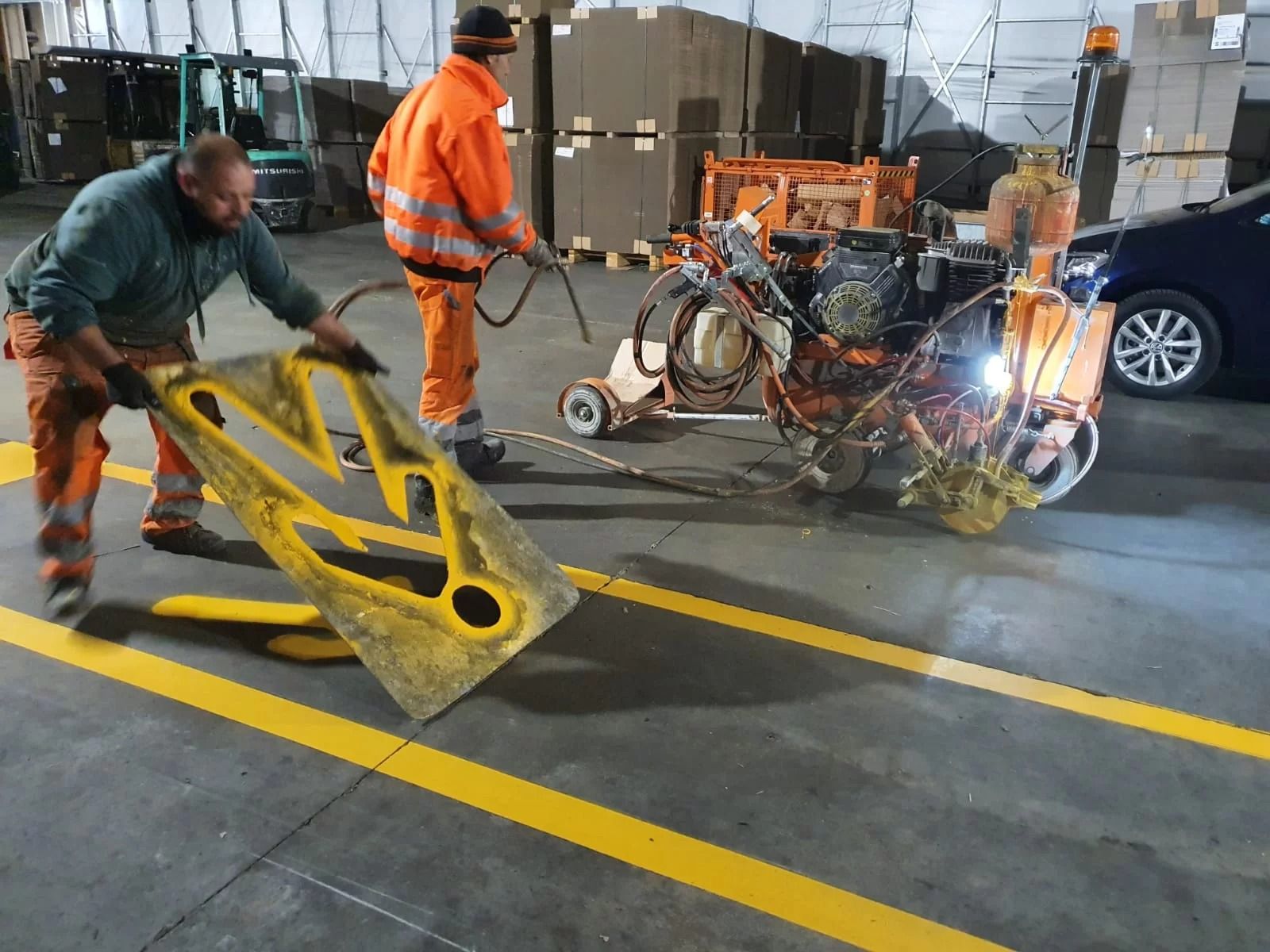 Due operai con indumenti arancioni ad alta visibilità dipingono le linee del pavimento utilizzando uno stencil e dei macchinari in un magazzino.