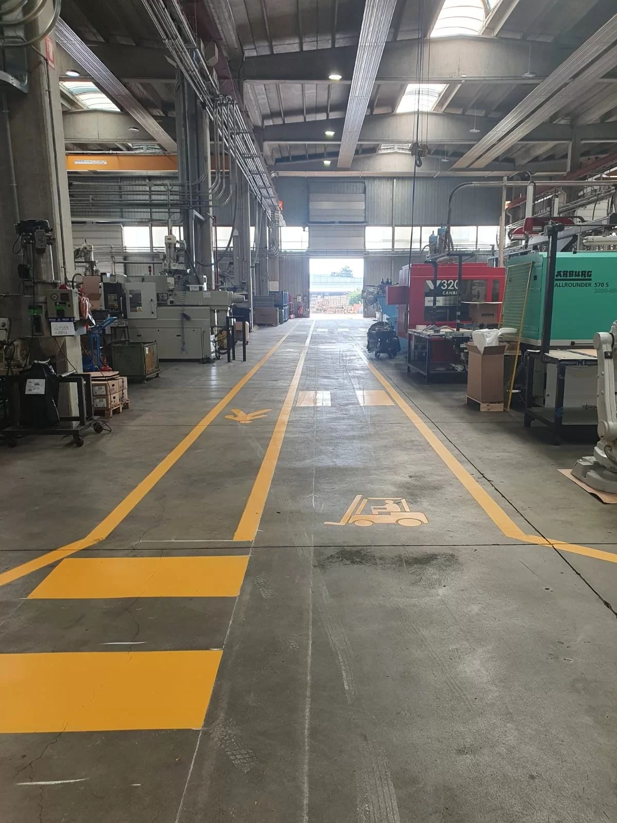 Area industriale con passerelle pedonali dipinte di giallo e simboli di sicurezza segnati sul percorso in cemento grigio.