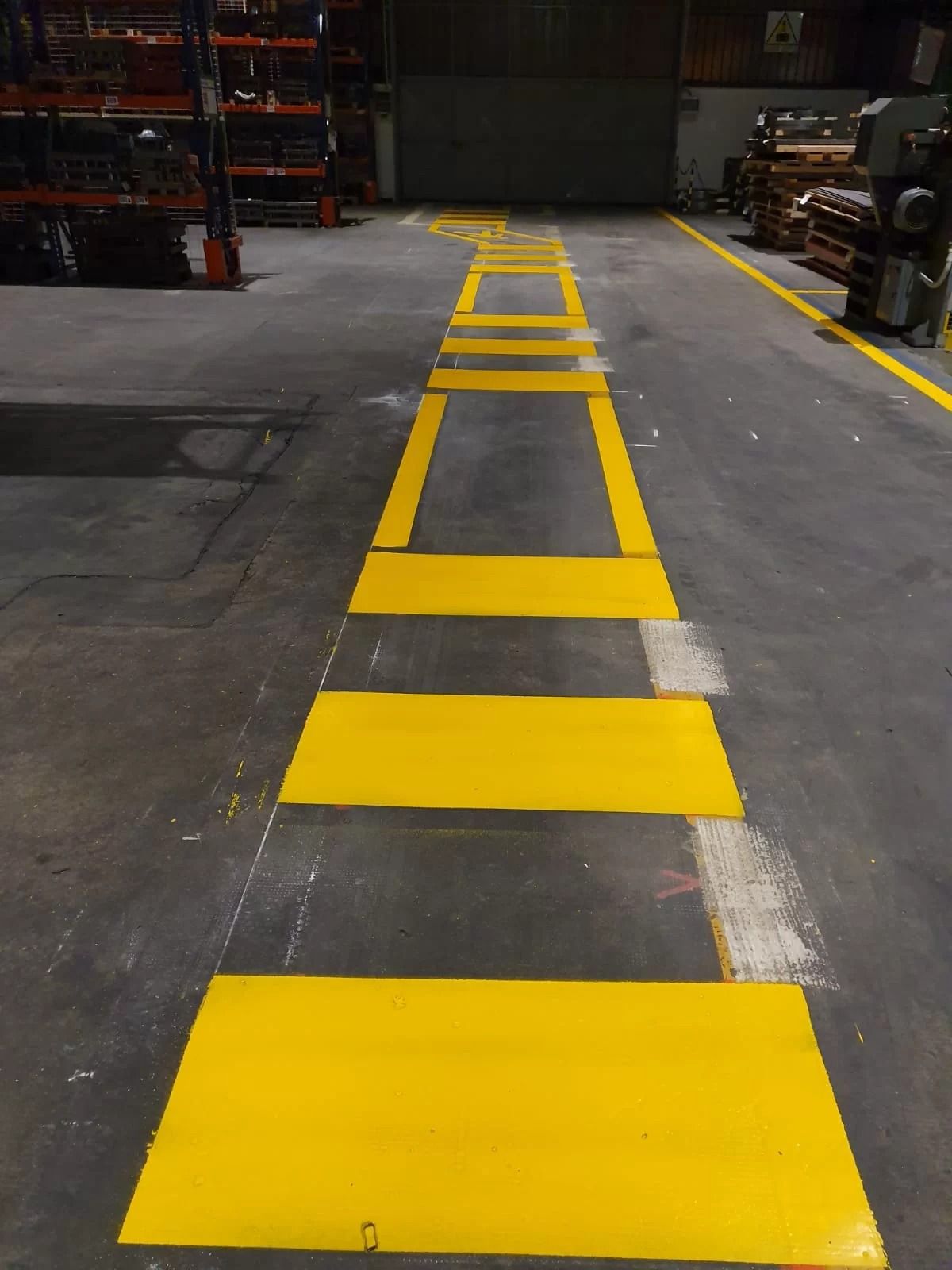 Un passaggio pedonale a livello del pavimento di un magazzino, segnalato da strisce giallo brillante e contorni rettangolari dipinti su cemento grigio.