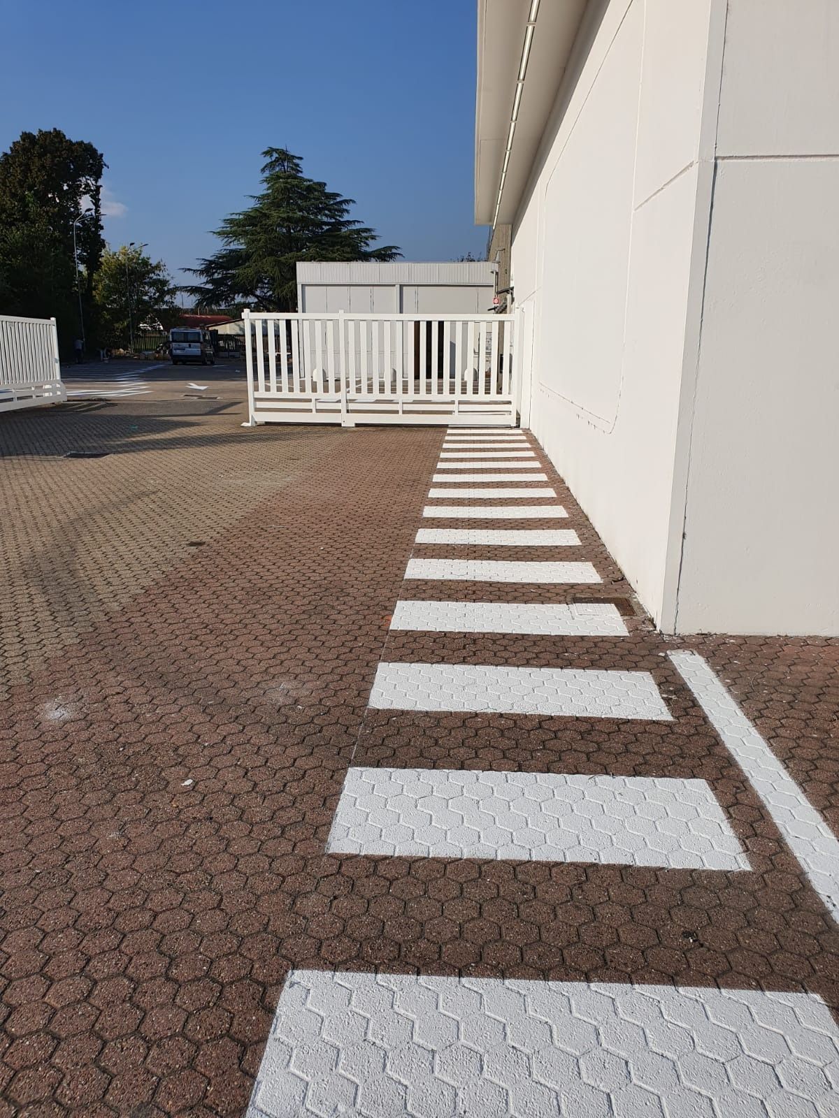 Un percorso pedonale delimitato da rettangoli dipinti di bianco su un parcheggio pavimentato in mattoni, accanto a un edificio bianco.