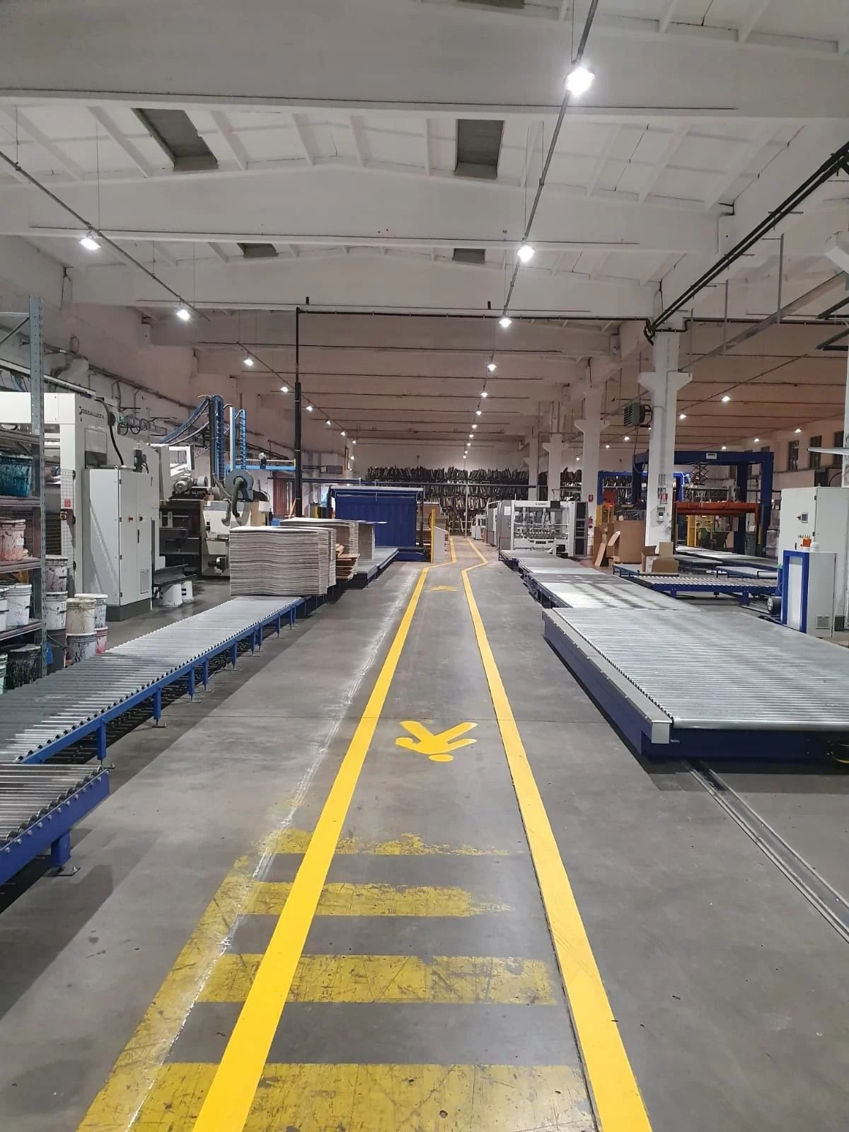 Un ampio corridoio di magazzino con segnaletica orizzontale gialla e segnali di attraversamento pedonale tra i sistemi di nastri trasportatori industriali.