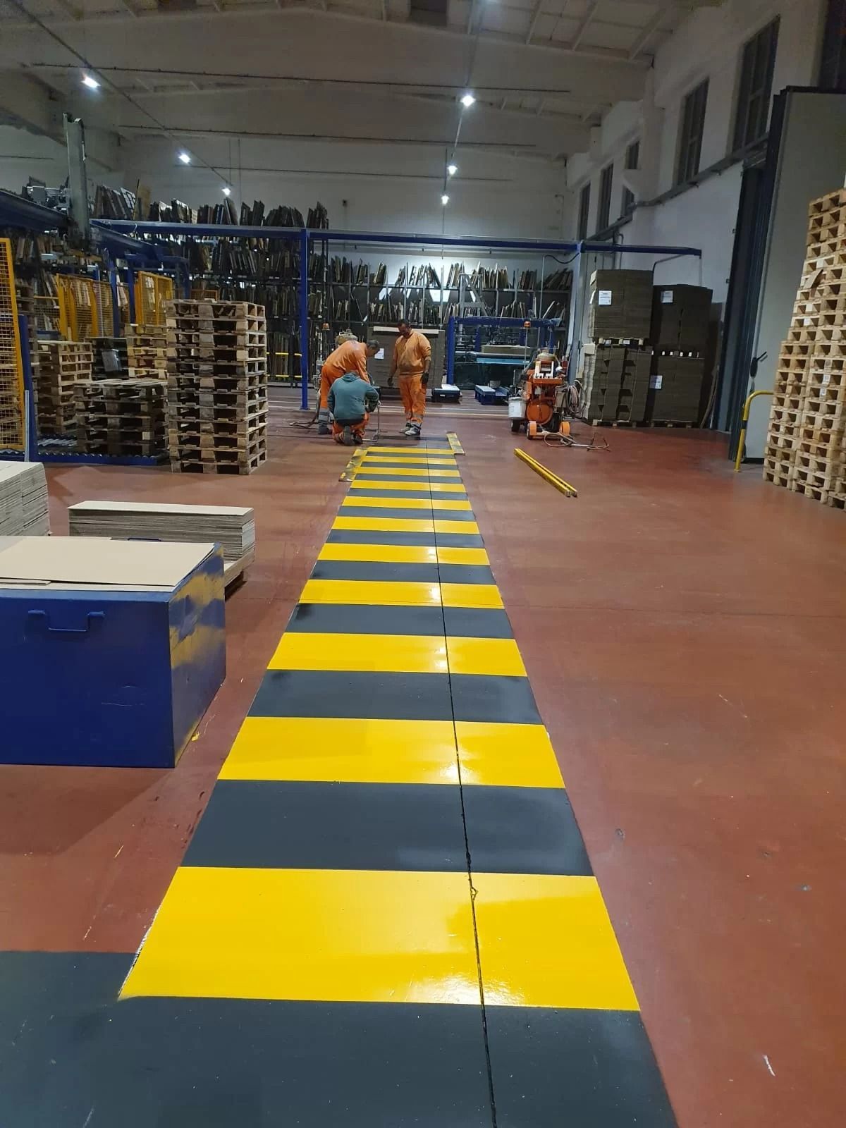 Un percorso pedonale a strisce gialle e nere segna un passaggio all'interno di un magazzino, con gli operai visibili sullo sfondo.
