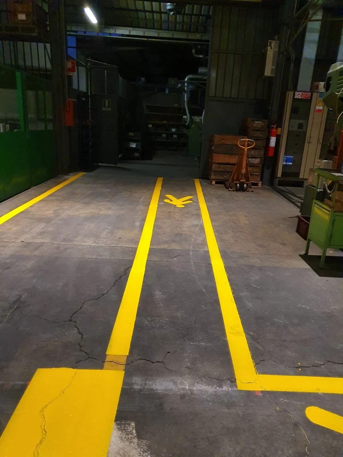 L'interno di una fabbrica è delimitato da linee di passaggio dipinte di giallo, una freccia direzionale e, sullo sfondo, un transpallet manuale.