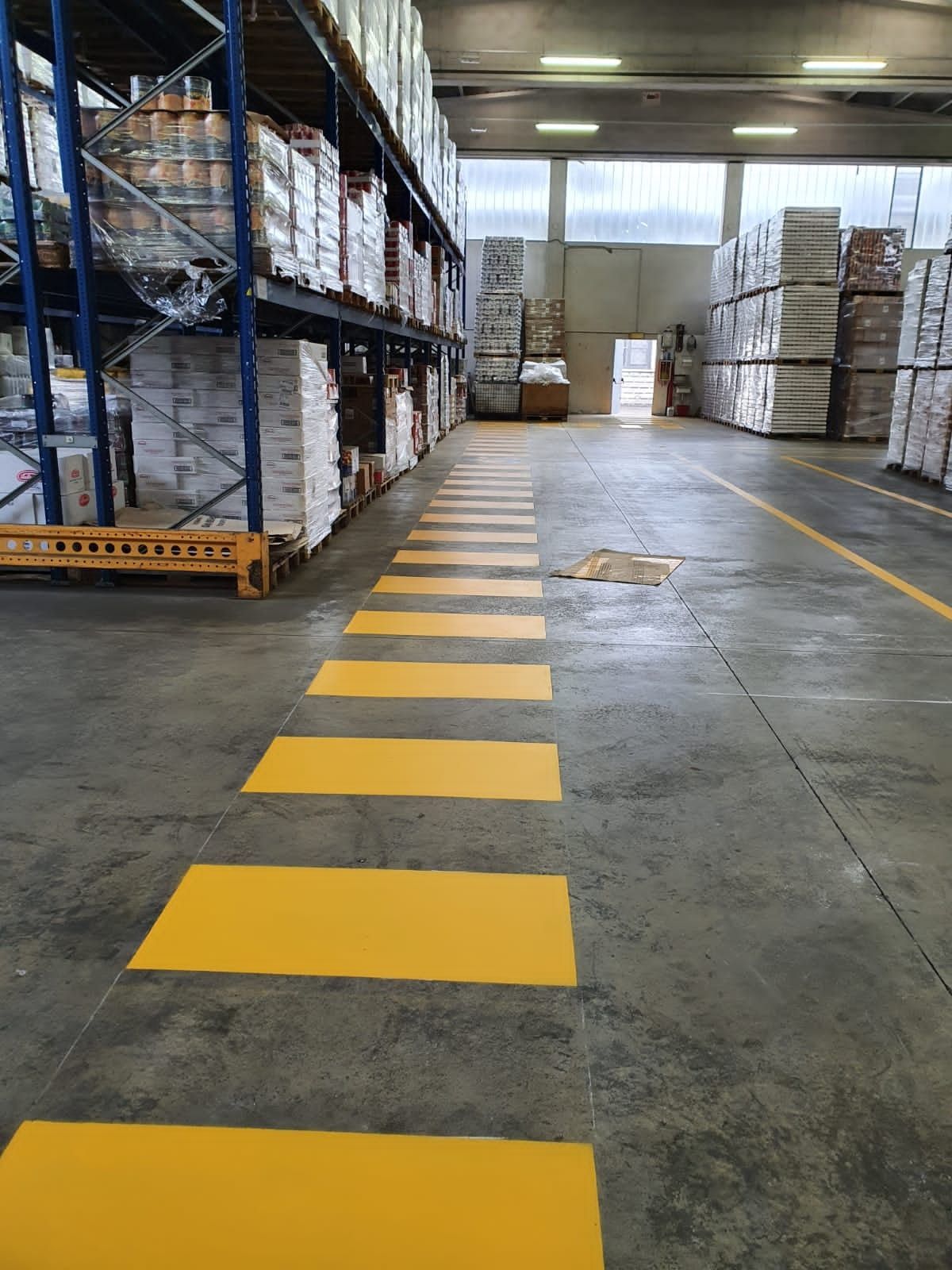 Una vista lungo un corridoio di un magazzino, con un attraversamento pedonale di colore giallo brillante dipinto sul pavimento di cemento.